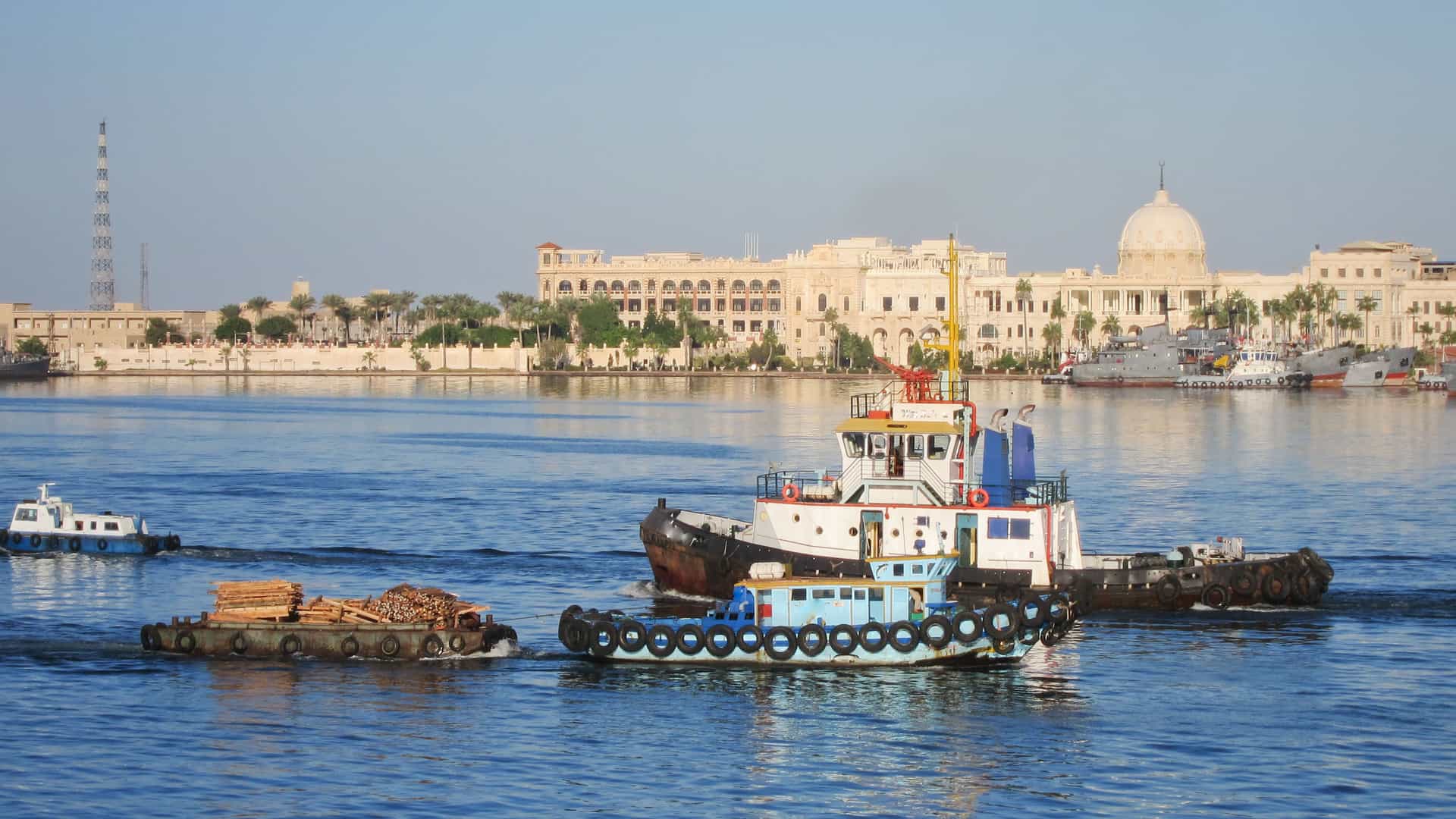 The harbor of Port Said, Egypt, a major port city at the northern end of the Suez Canal, a man-made waterway that connects to the Nile River system. In the foreground, several tugboats and barges are navigating the calm, blue water. One barge is loaded with large pieces of wood. On the far bank, a line of light-colored, ornate buildings with domes and arched windows stands prominently. The background also shows industrial and port structures. The image captures the busy maritime and urban life of a significant city in the Egyptian region.