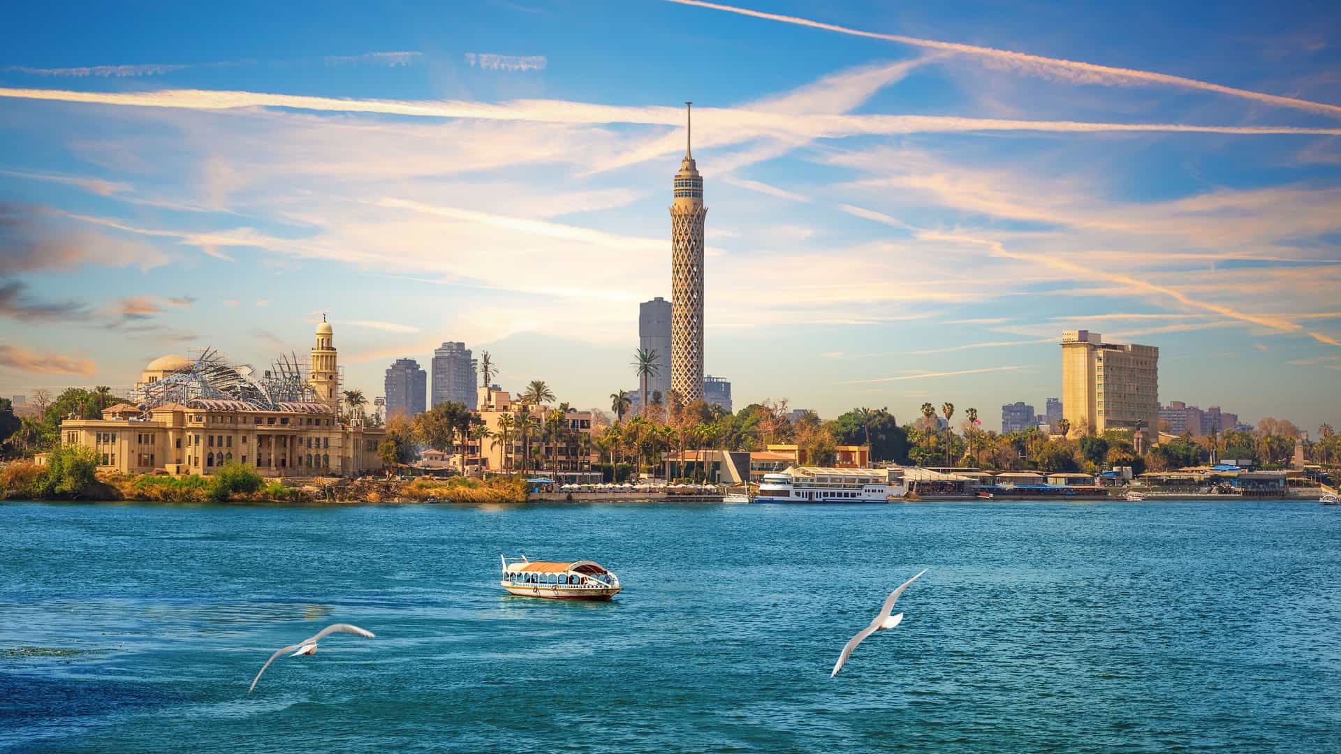 A beautiful wide shot of the Nile River in Cairo, Egypt, with the city's skyline in the background. The most prominent landmark is the towering Cairo Tower with its lattice design. The river is a deep turquoise, with a small boat floating in the foreground and a long river cruise ship docked on the far bank. Two seagulls are in mid-flight over the water. The sky is a dramatic mix of blue with white and orange clouds, illuminated by the late afternoon sun. The image captures the dynamic and historic cityscape of Cairo along the life-giving Nile River.