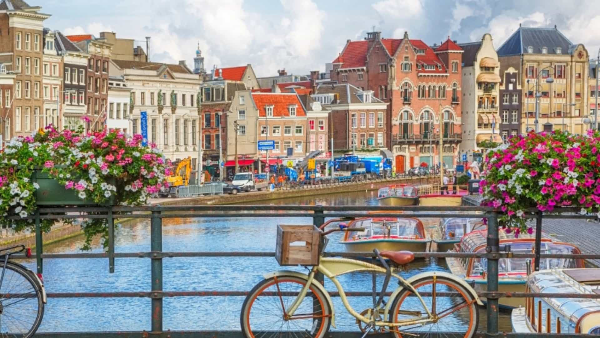 A quintessential scene of Amsterdam, Netherlands, a major hub for European river cruises. In the foreground, two bicycles are parked on a stone bridge railing adorned with large planters overflowing with vibrant pink and white flowers. Below, a calm canal is filled with various boats and barges. The background is a colorful line of traditional Amsterdam buildings with their iconic gabled roofs, showcasing the charming and unique cityscape of this European region.
