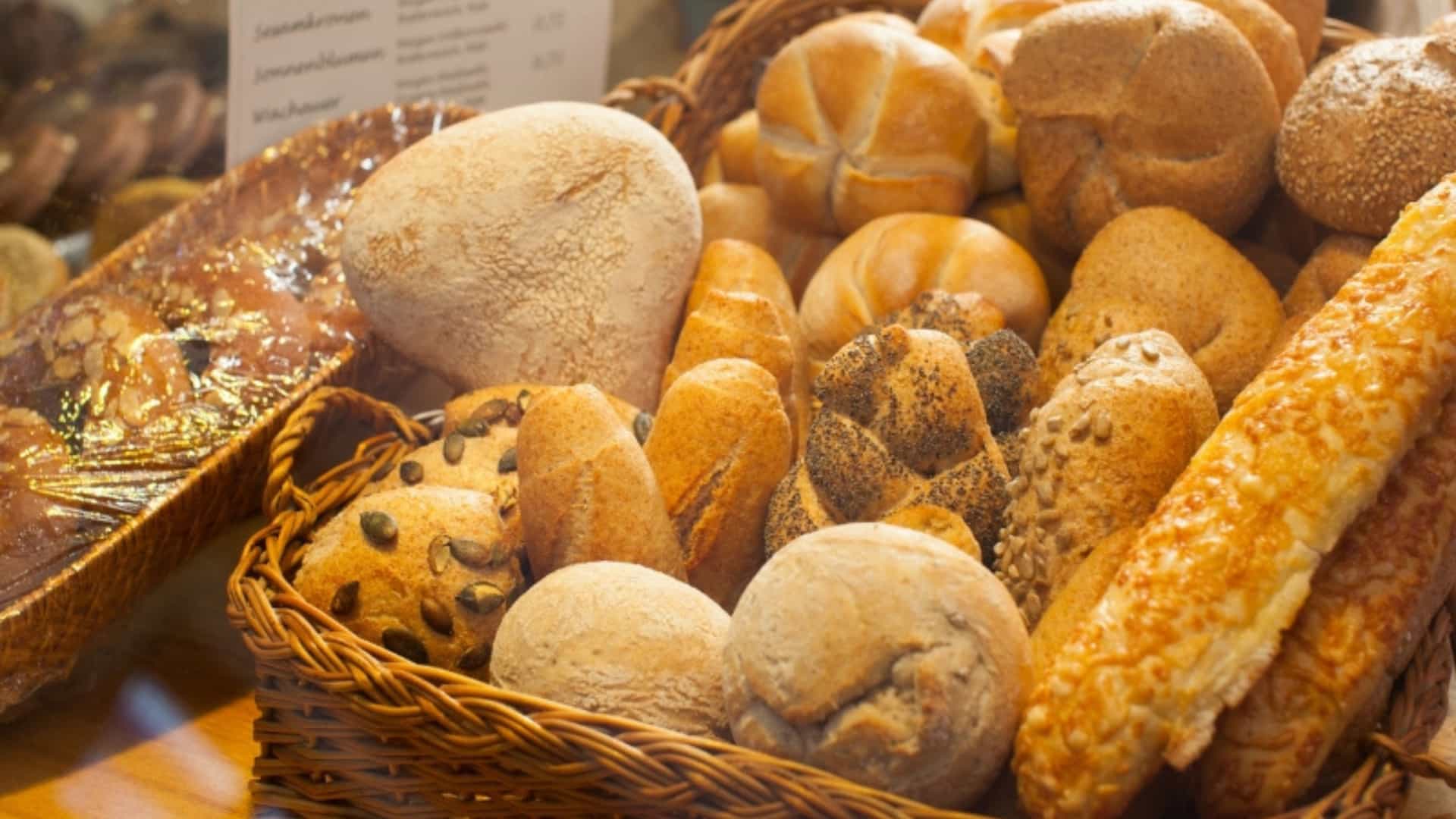 A delicious assortment of fresh Austrian bread rolls and pastries is displayed in a woven basket inside a bakery window. The basket is filled with a variety of baked goods, some with smooth tops, some dusted with seeds like poppy and sesame, and others with a braided texture. The warm, golden-brown colors of the bread are inviting. This image represents the local culinary delights of the European region, particularly Austria, a common stop on many river cruises.