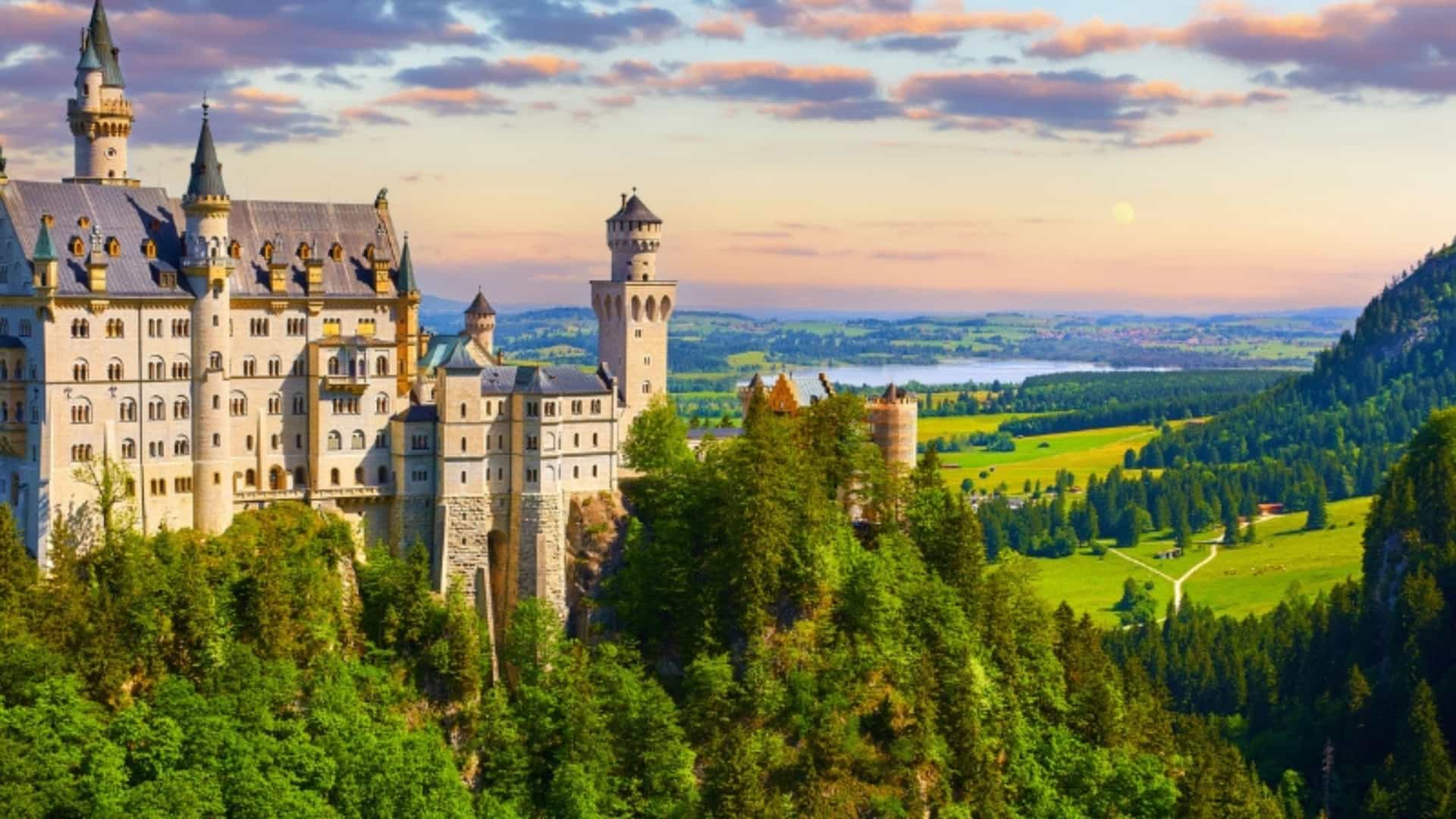 An iconic view of Neuschwanstein Castle, a famous fairytale-like fortress in Bavaria, Germany, a region linked to European river cruises. The castle, with its white walls, turrets, and towers, is perched on a rocky cliff and surrounded by a dense green forest. In the distance, a serene lake and lush green valleys stretch out toward the horizon. The sky is a dramatic mix of orange, pink, and purple clouds, with a soft moon or sun in the distance, creating a magical atmosphere. This image captures one of the most famous landmarks in the European region.