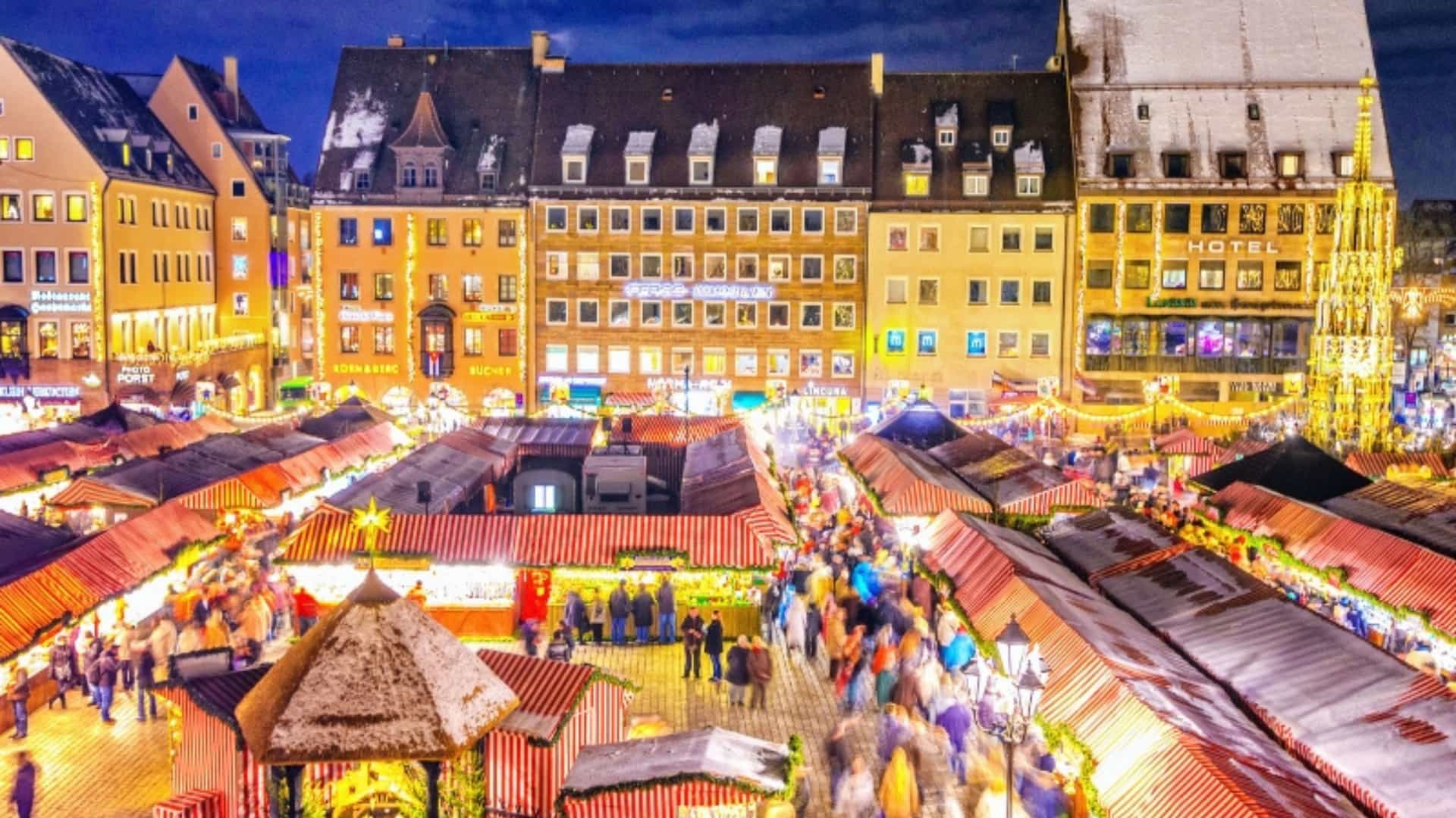 A beautiful aerial view of the Nuremberg Christmas market in Germany, a popular seasonal event in the European river cruise region. The marketplace is filled with rows of small, festive stalls with red-and-white striped roofs. A large crowd of people, shown as blurred streaks of color, moves through the aisles. The scene is illuminated by countless warm, glowing lights and a large, decorated Christmas tree. The historic buildings in the background are also lit up and dusted with a light layer of snow, creating a magical winter atmosphere.