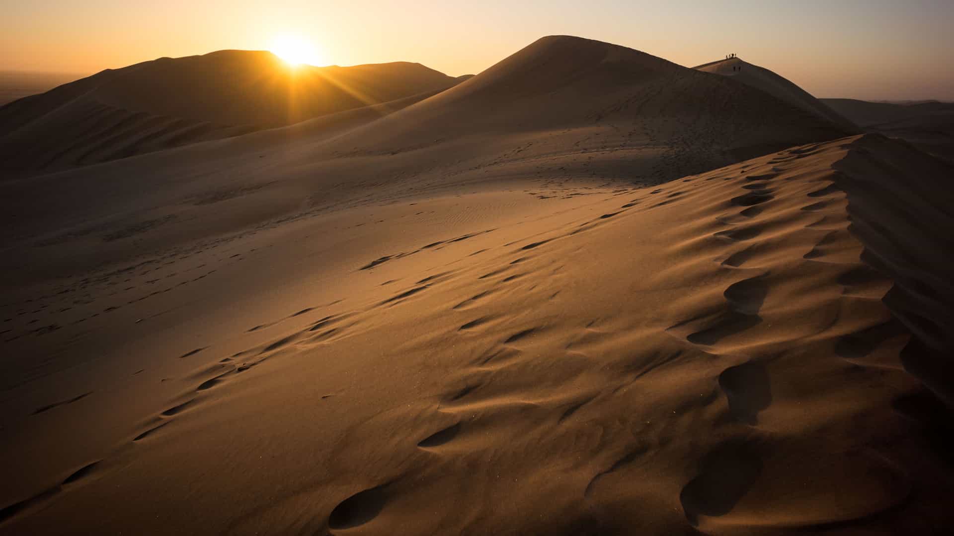 A stunning photograph of the desert dunes near Walvis Bay, Namibia, at sunset. The sun, a bright golden orb, is setting behind a distant dune, casting a warm, orange glow across the landscape. The undulating sand dunes are covered in footprints and show intricate patterns created by the wind. A distant figure is visible on a far dune, providing a sense of scale. The image captures the serene and vast beauty of the desert landscape in the Namibia region.
