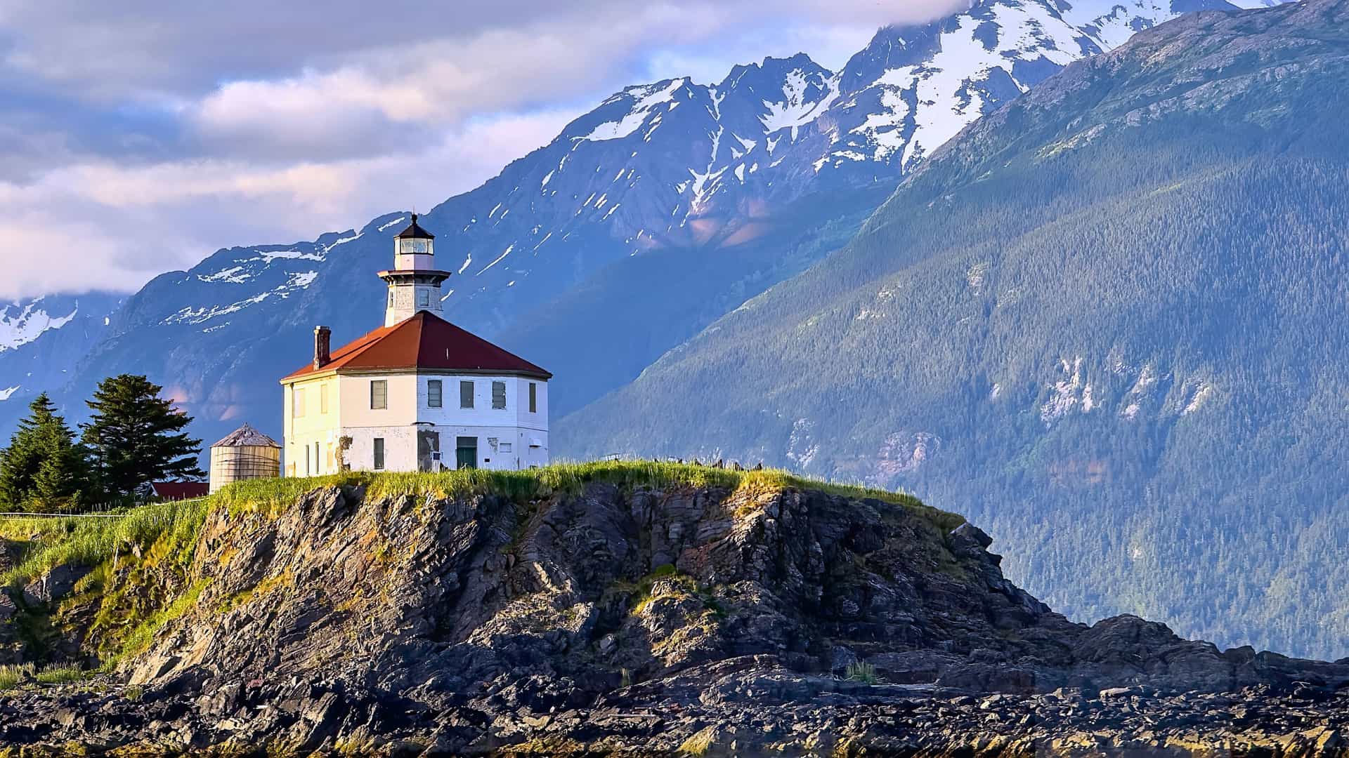 Explora journey view of a historic lighthouse perched on a rocky cliff against a backdrop of snow-capped mountains along the U.S. Pacific Coast.