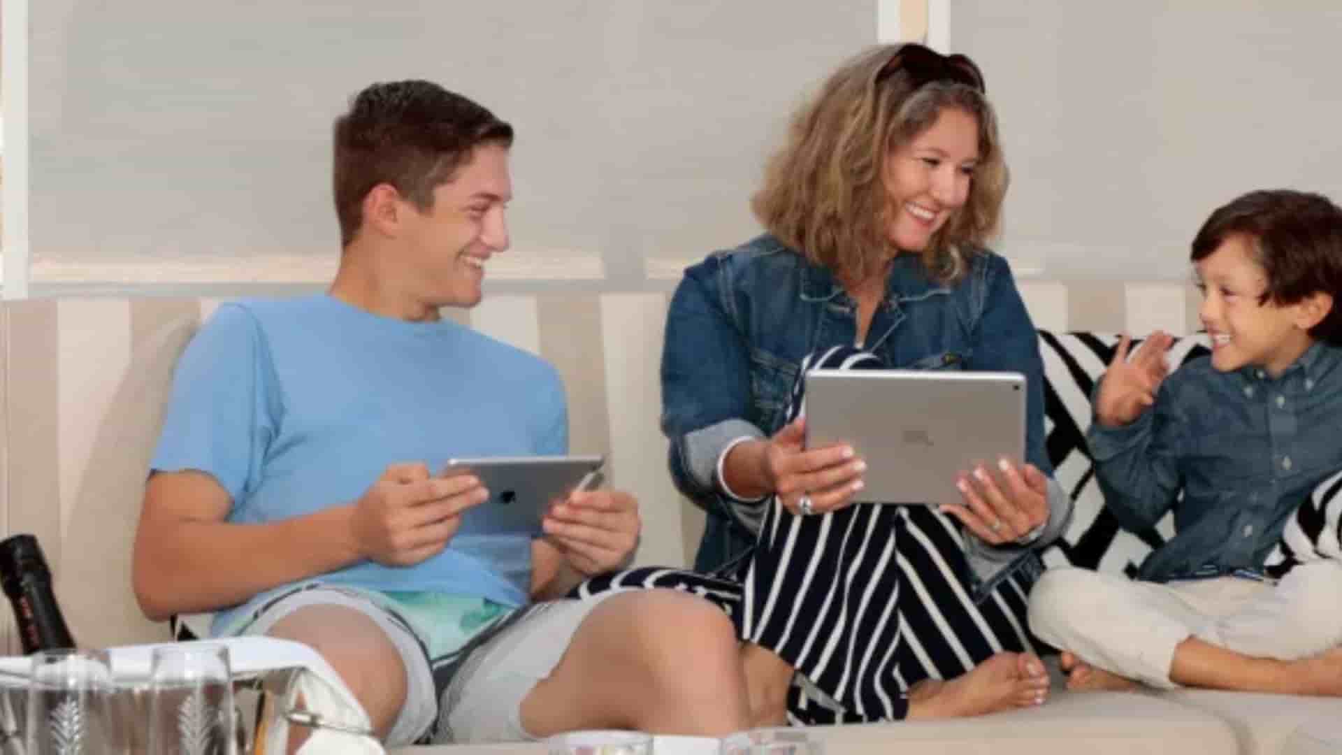 A family using iPad tablets on a couch aboard a cruise ship with a bucket of iced drinks in the foreground.