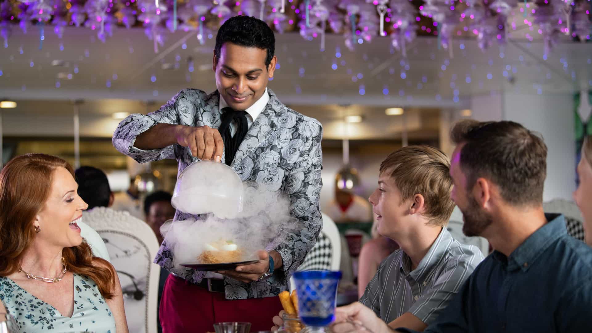 Waiter lifting up a smoke-filled cloche for a family's dining table at Wonderland on a Royal Caribbean ship.