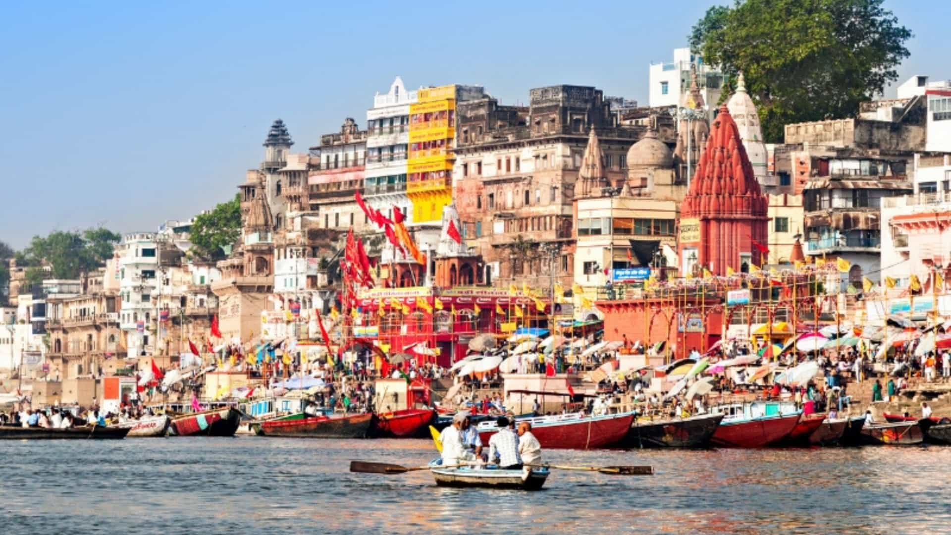 A vibrant and bustling scene along the banks of the Ganges River in Varanasi, India. The image is taken from the water, showing the city's famous ghats, or steps leading down to the river. Colorful, ancient buildings and temples rise up the hillside, with a large crowd of people and boats lining the banks. A small rowboat with people in it is in the foreground. The bright colors, clear blue sky, and lively activity capture the spiritual and cultural significance of the Ganges River in the India region.