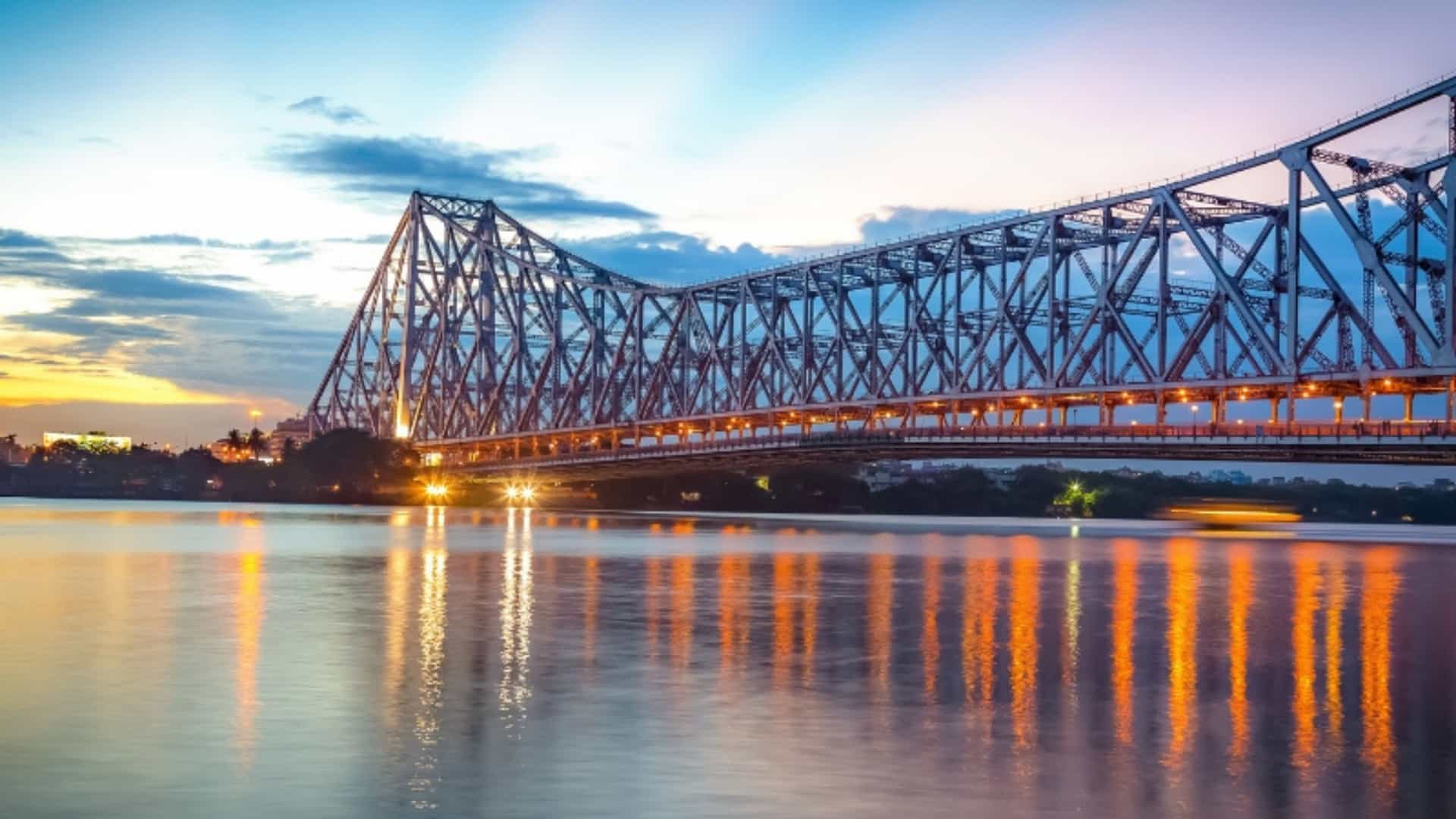 A beautiful sunset view of the Howrah Bridge over the Hooghly River in Kolkata, India. The massive cantilever bridge is illuminated, with its lights reflecting on the calm water below. The sky above is a blend of deep blue, orange, and purple hues, with light rays streaking from the horizon. The silhouette of buildings and trees is visible on the far bank. The image captures an iconic landmark in the Ganges River region.