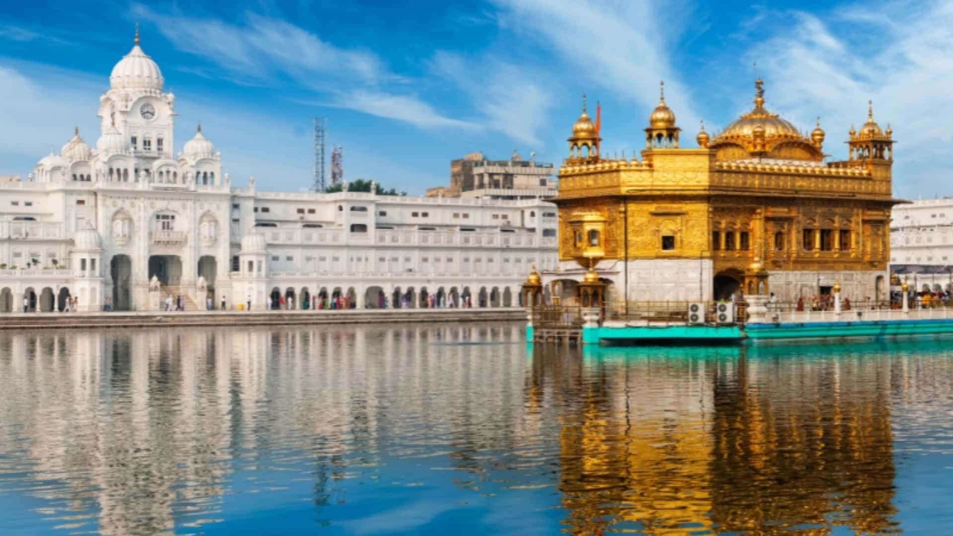 A serene view of the Golden Temple, also known as Sri Harmandir Sahib, in Amritsar, Punjab, India. The magnificent, gold-plated temple stands on a small island in the center of the Amrit Sarovar, a large sacred pool. The golden structure reflects beautifully on the clear, blue water. A walkway connects it to the surrounding marble complex, which includes other white-domed buildings. The scene, under a bright blue sky with wispy clouds, captures the peaceful and spiritual essence of this important Sikh landmark in the Ganges River region.