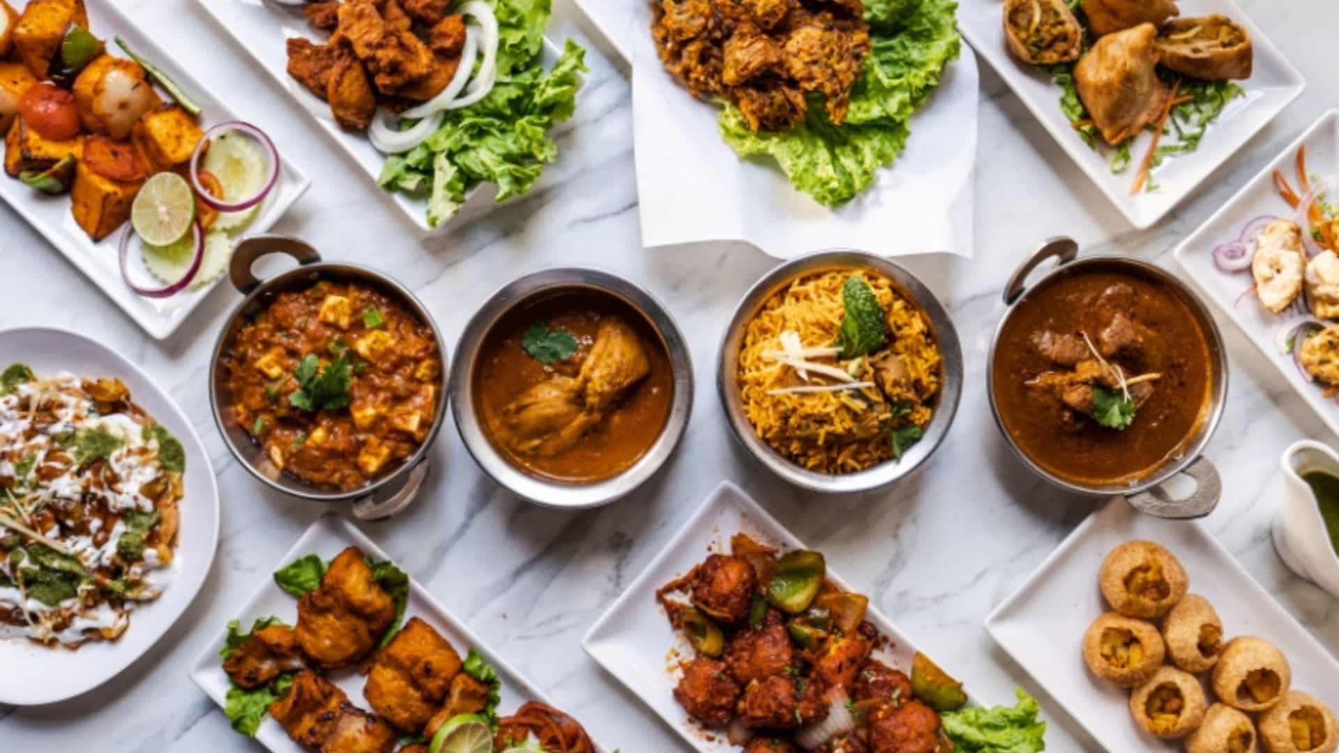 An overhead view of a diverse selection of traditional Indian dishes, representing the cuisine of the Ganges River region. The spread includes various curries, platters of fried snacks like pakoras and samosas, a bowl of biryani rice, and small puri balls. The food is arranged on white plates and in metal bowls on a light-colored marble surface. The vibrant colors and textures of the dishes highlight the rich and flavorful culinary traditions of India.