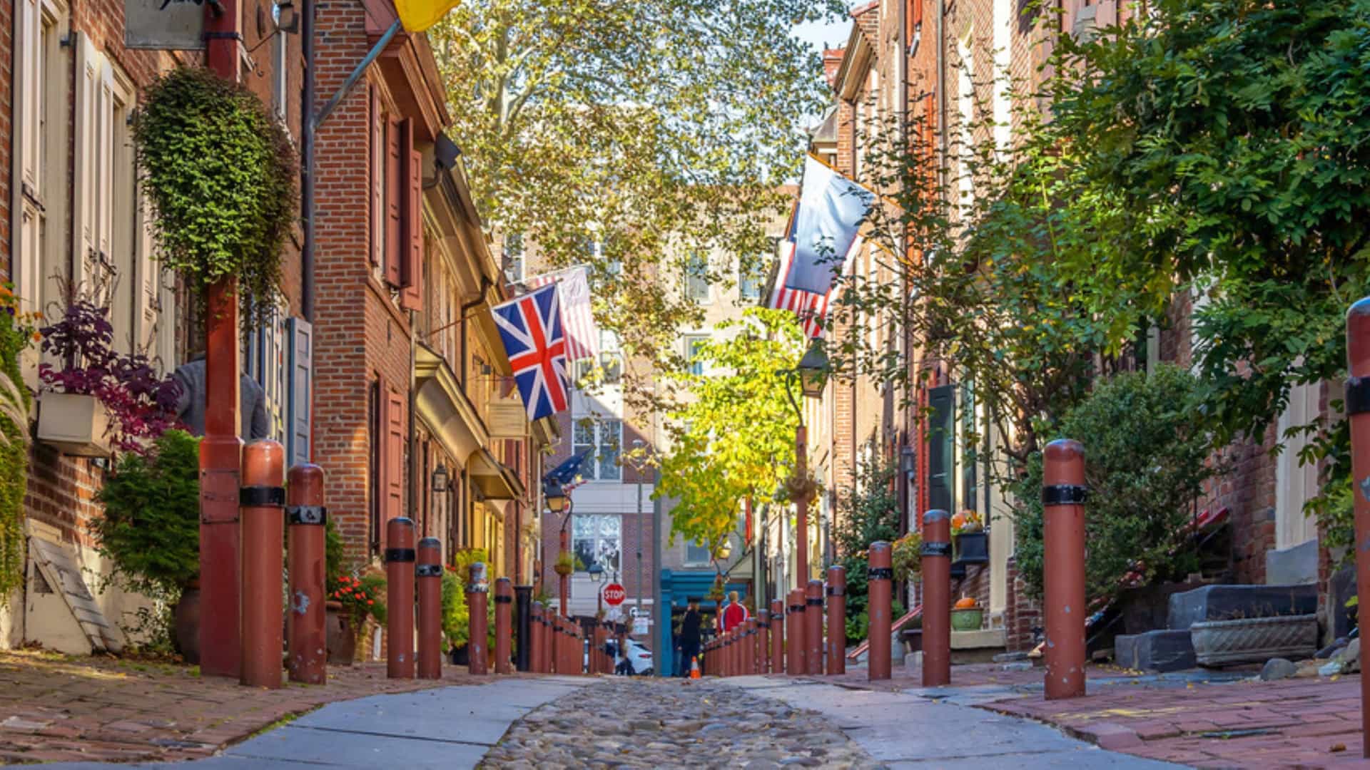 "A low-angle view of Elfreth's Alley in Philadelphia, featuring historic brick row houses and a cobblestone street. Early American and British flags hang from the buildings under a canopy of green trees, capturing the colonial atmosphere of America's oldest continuously inhabited street.