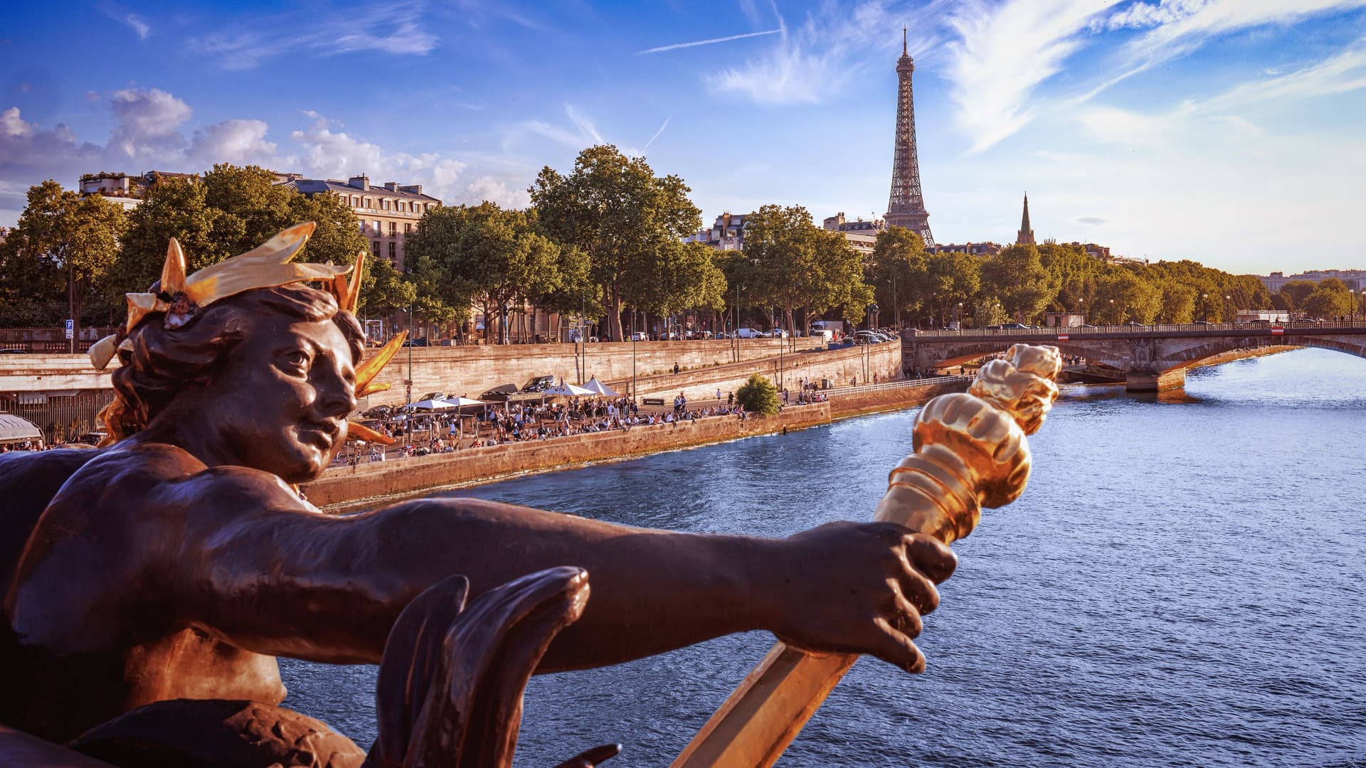 A gilded bronze sculpture on the Pont Alexandre III bridge extends over the Seine River in Paris, with the Eiffel Tower rising above tree-lined banks on a sunny afternoon..