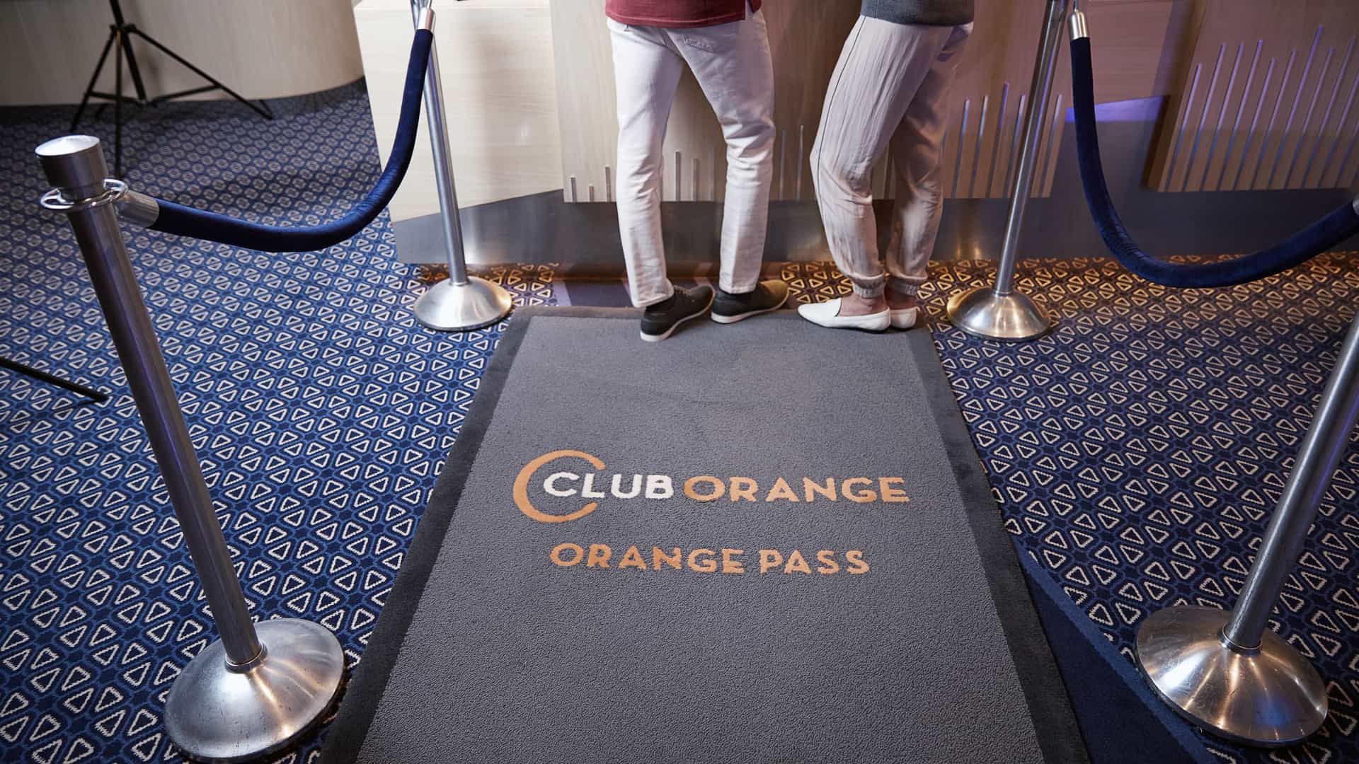 Guests entering the Holland America Club Orange priority access entrance, marked by a Club Orange Orange Pass mat and blue rope stanchions.