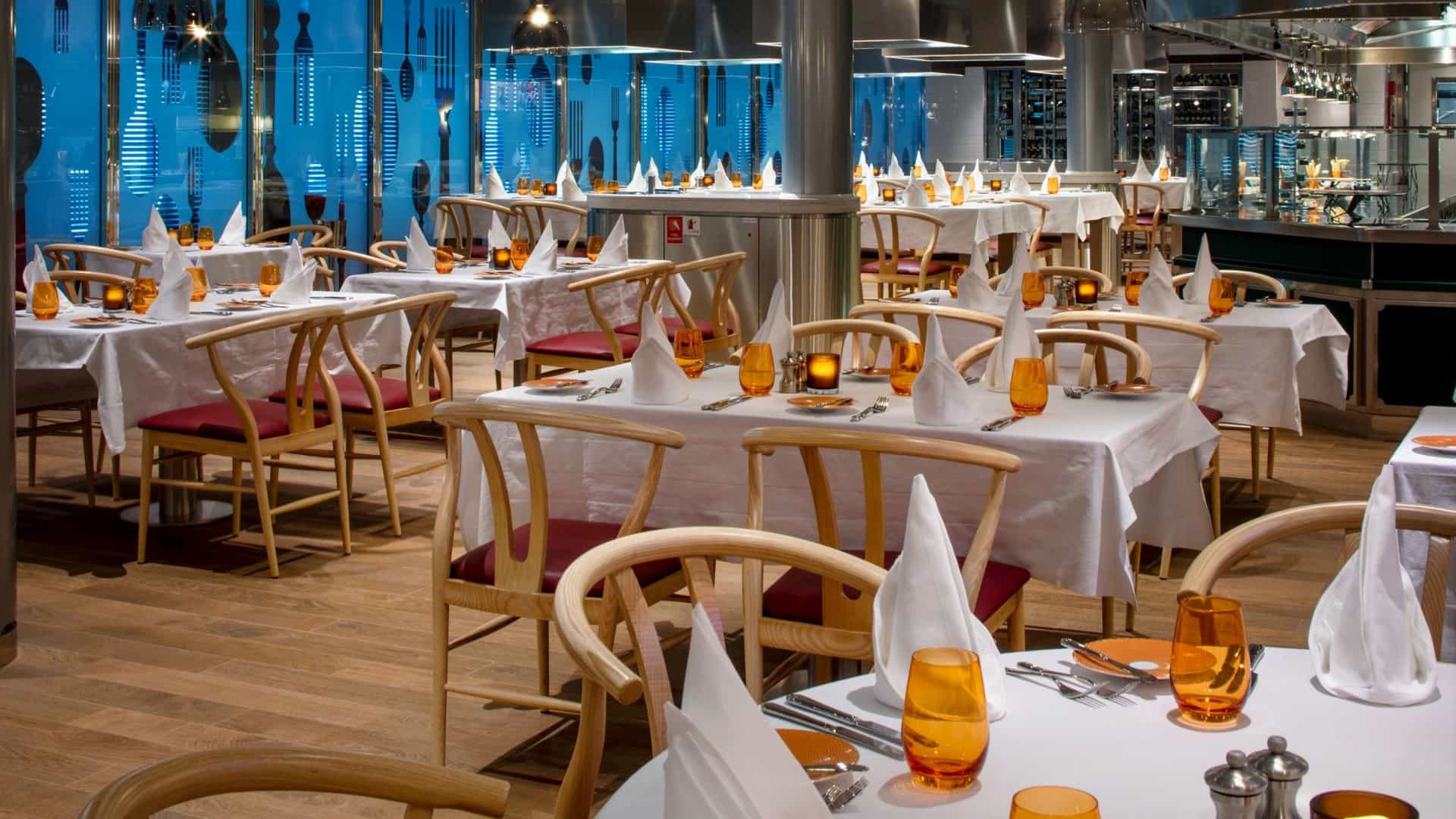 The elegantly set main dining room aboard a Holland America ship featuring white linen tables, light wood chairs, amber glassware, and panoramic ocean views through floor-to-ceiling windows.