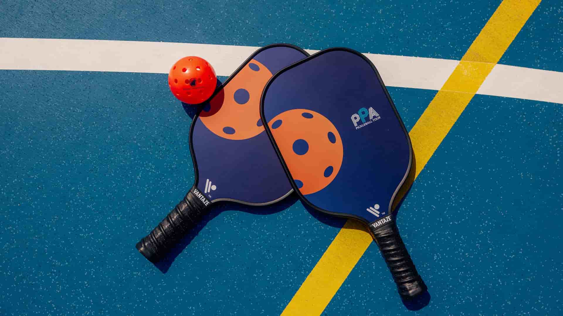 Two branded Vantaze pickleball paddles crossed on a blue court surface with white and yellow lines alongside a red Holland America Line pickleball.