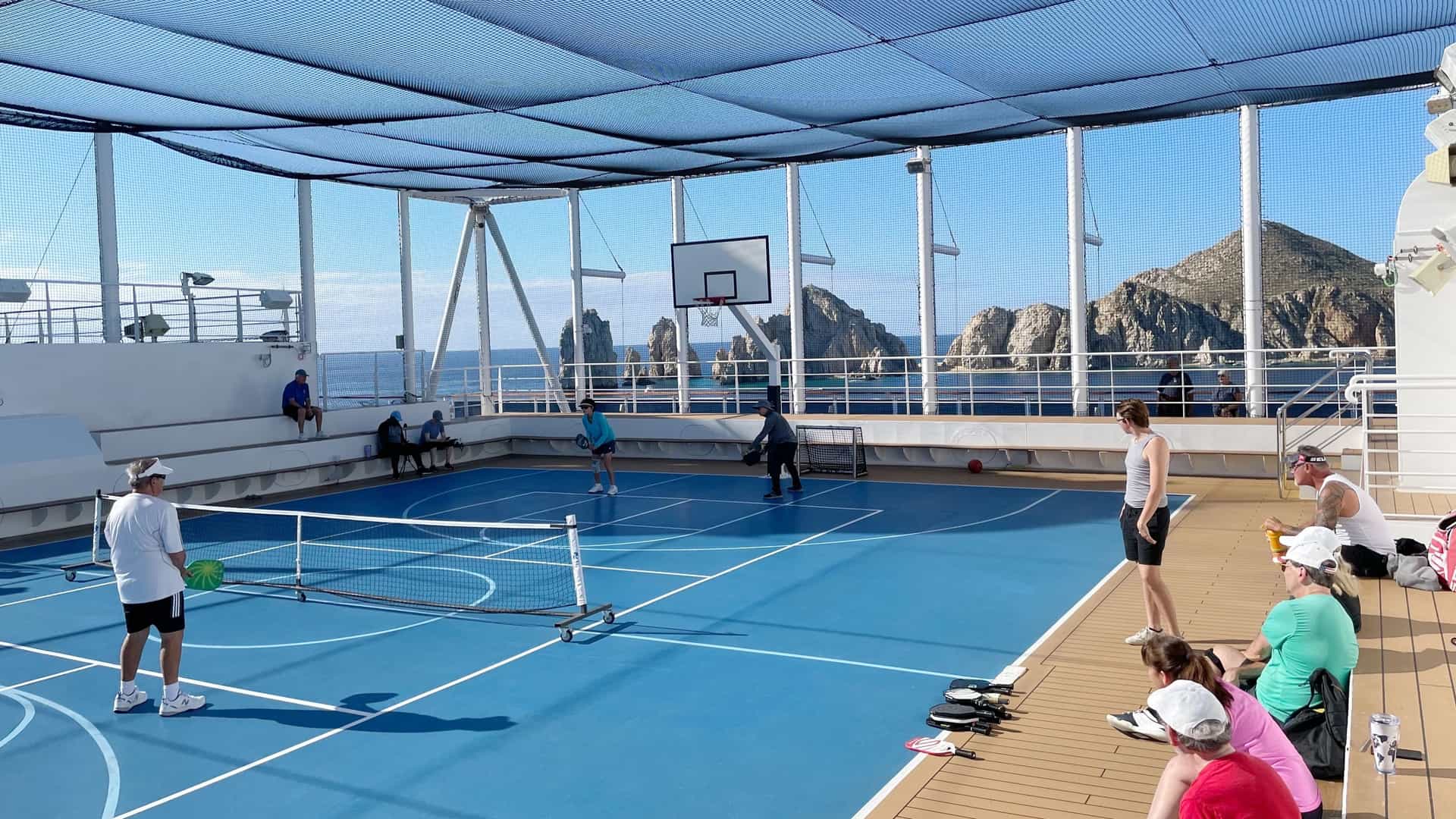 Passengers play a pickleball match on an outdoor sports court aboard a Holland America Line cruise ship, with spectators watching and the rock formations of Cabo San Lucas, Mexico, visible in the background.