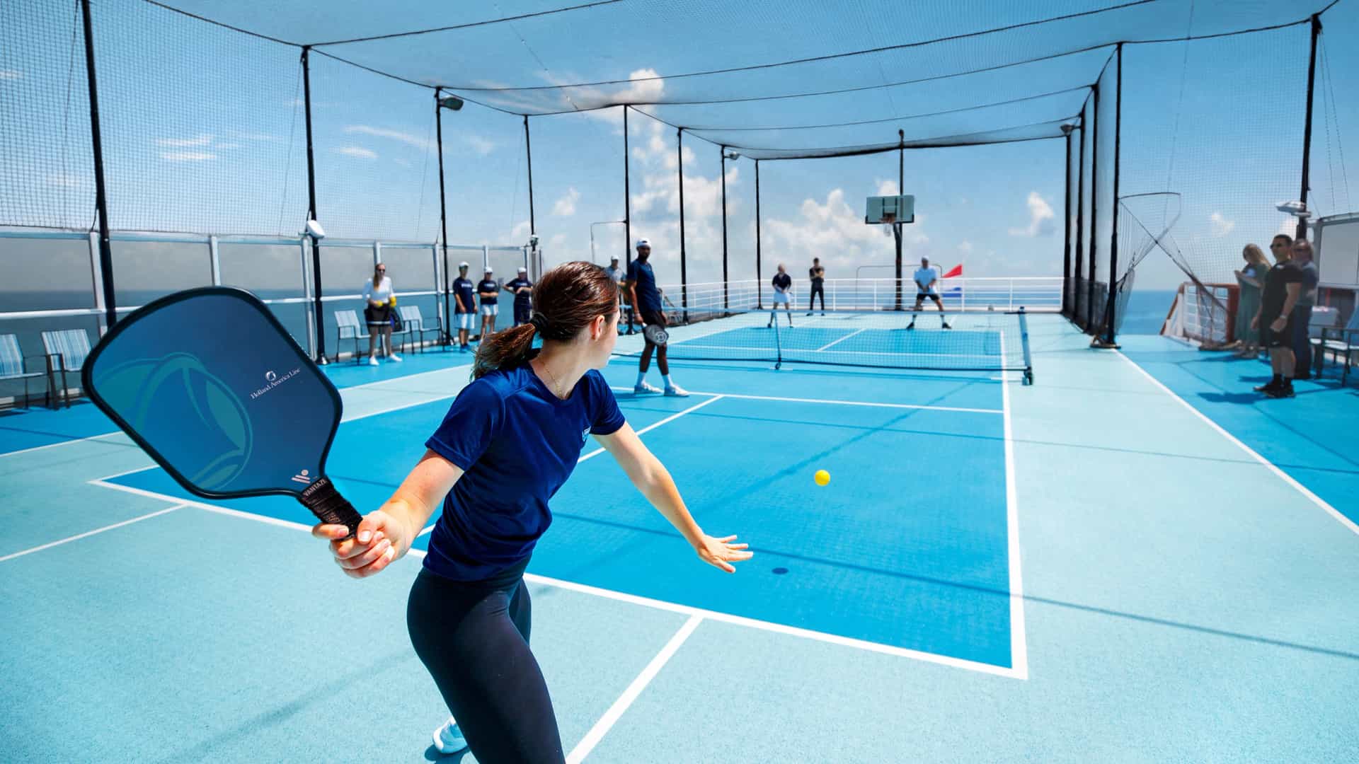 Woman mid-rally on Holland America Line's top-deck pickleball court with ocean views and fellow guests watching.