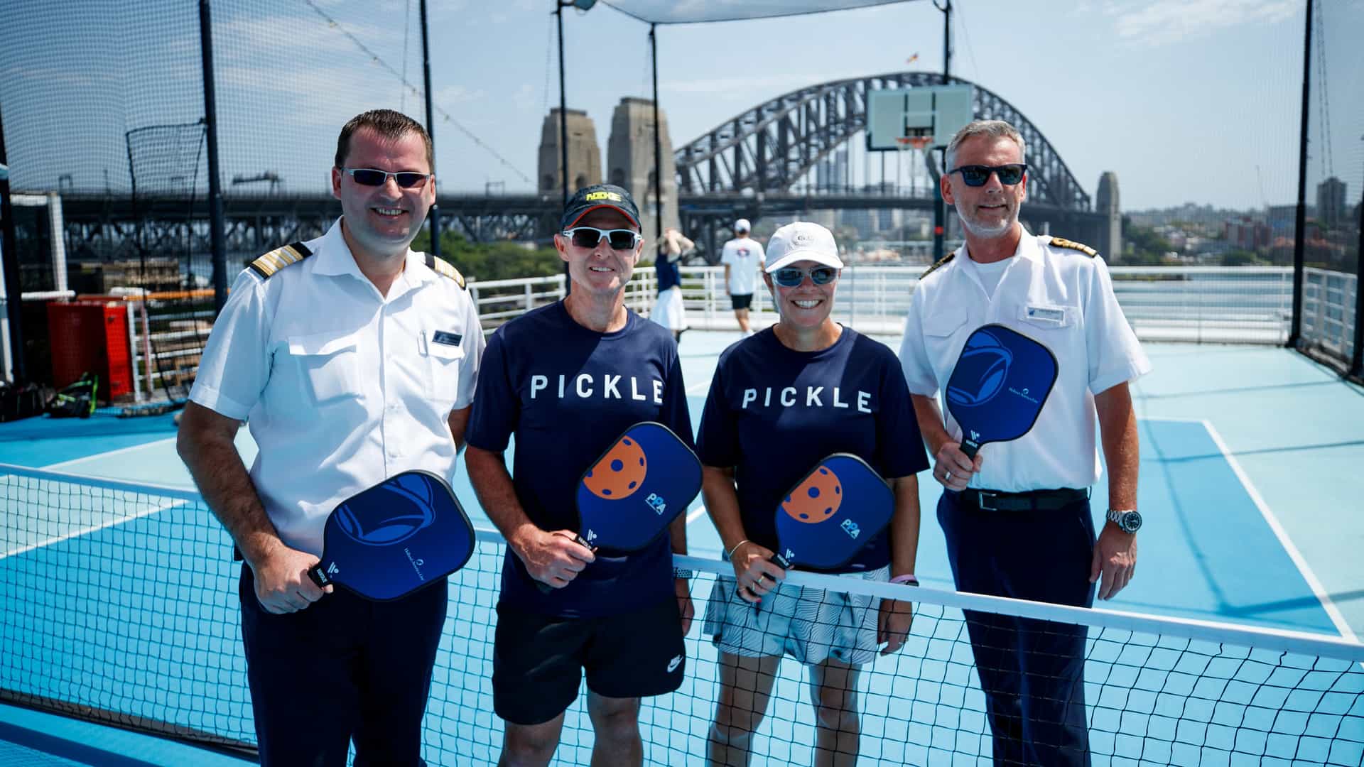 Holland America Line officers and guests posing with PPA pickleball paddles on a top-deck court with the Sydney Harbour Bridge in the background