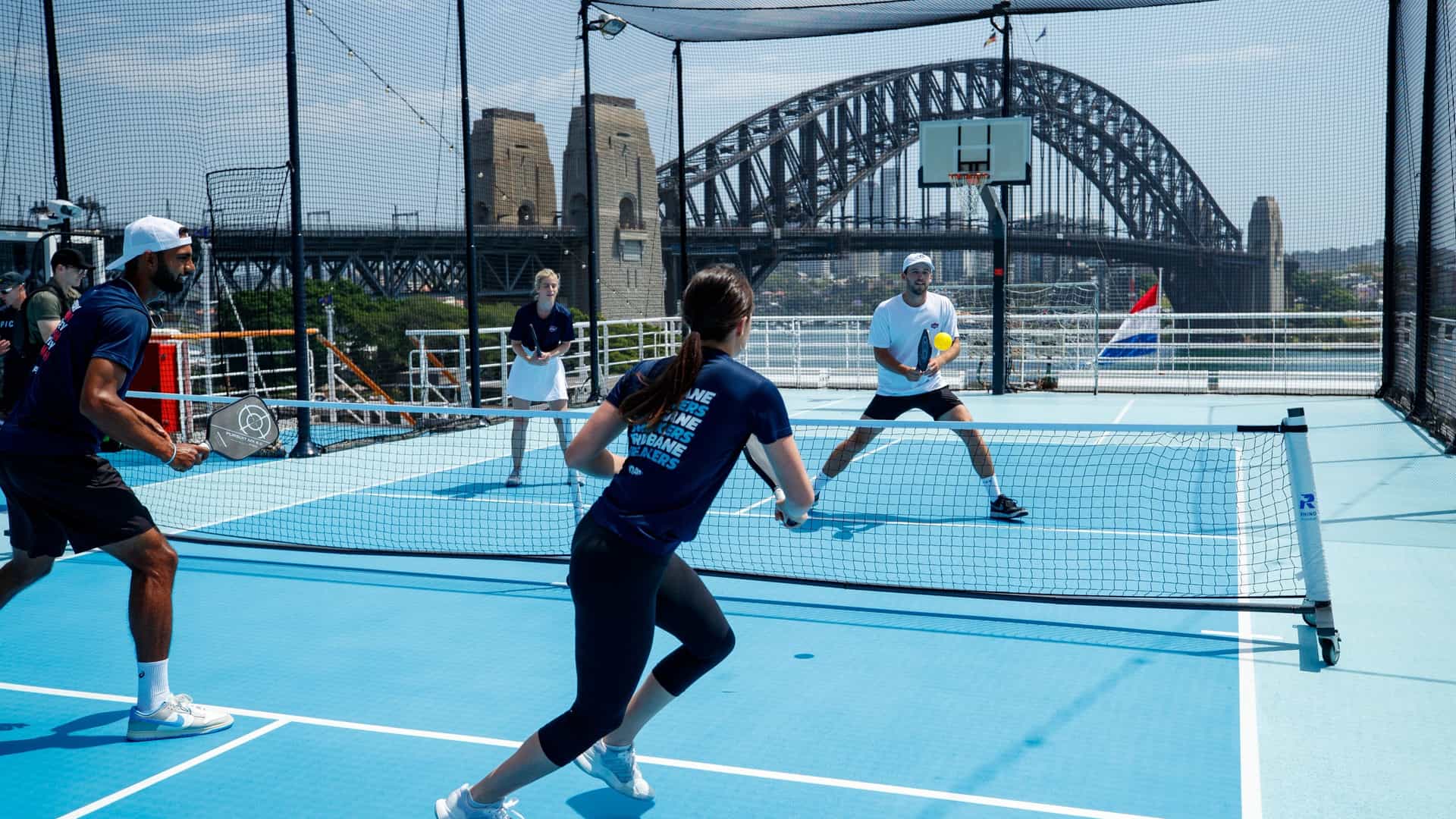 Passengers play a doubles pickleball match on an outdoor blue sports court aboard a Holland America Line cruise ship, with the iconic Sydney Harbour Bridge in the background.
