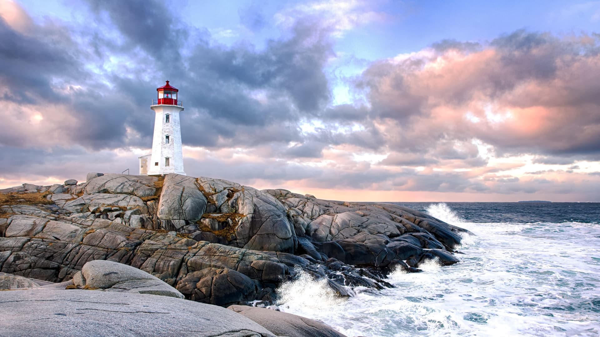 Peggy's Cove Lighthouse stands on rocky granite shore with waves crashing against rocks under a dramatic cloudy sky at sunset.