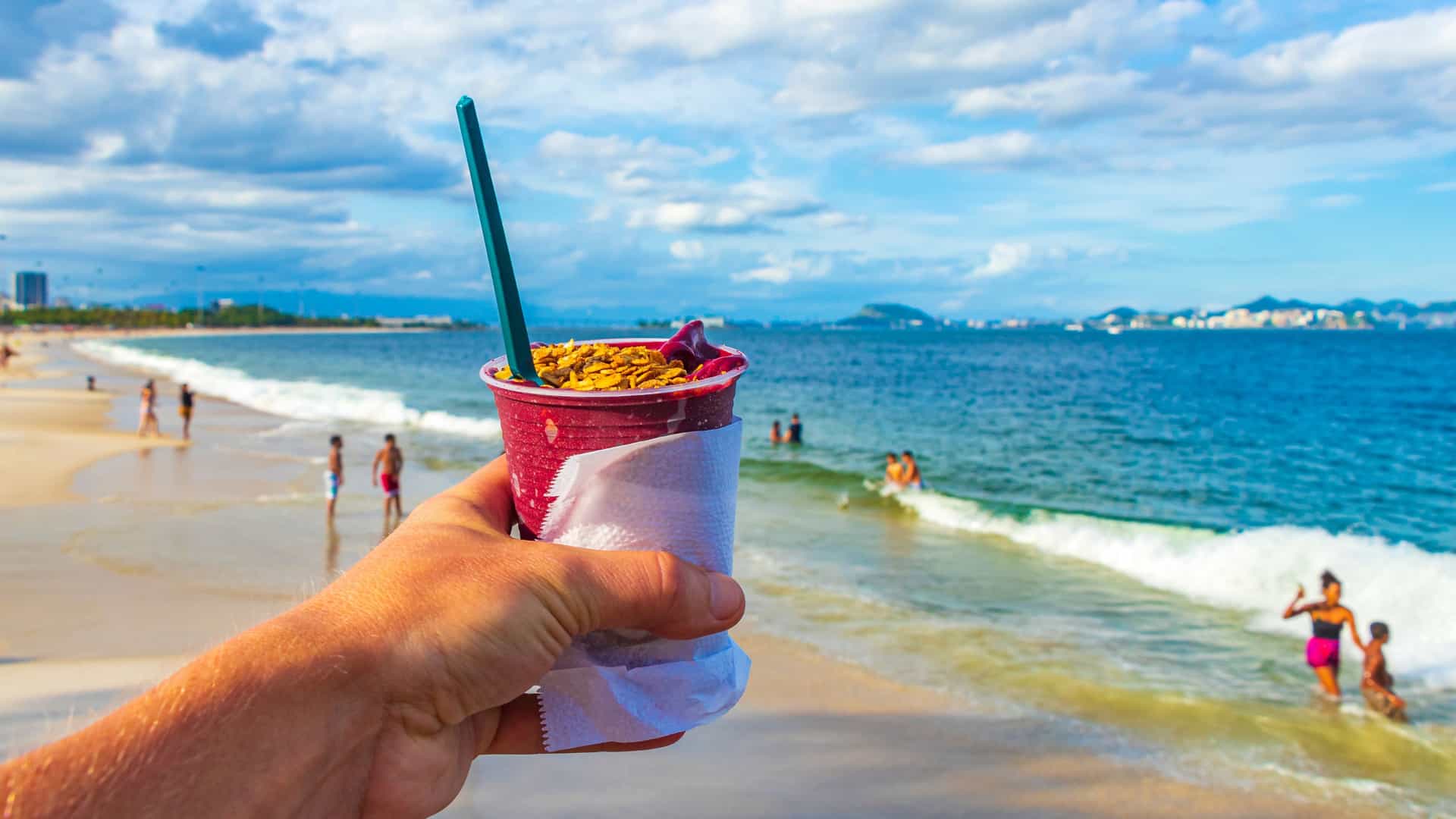 Image of a handing holing up a cup of acai sorbet on the beach with the shore in the background.