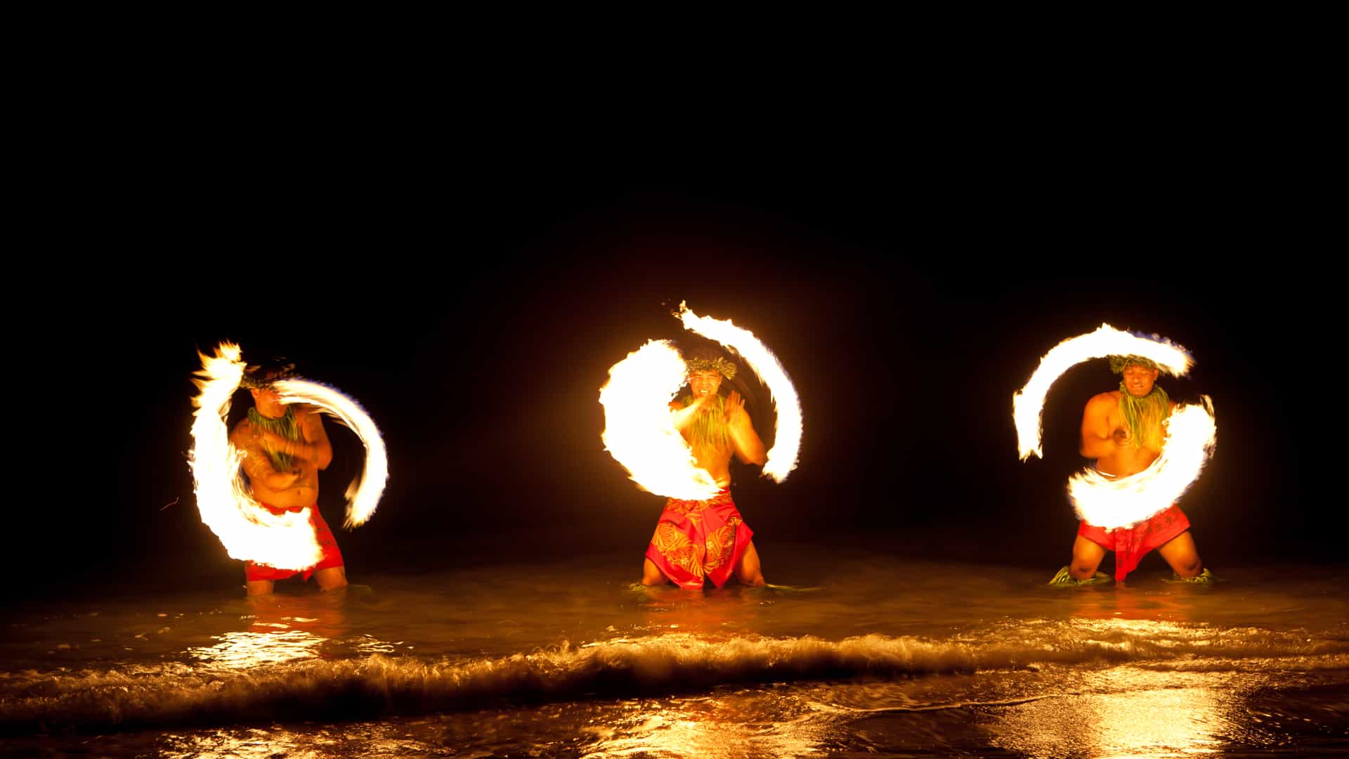 Three Hawaiian dancers perform with fire in the water, creating a vibrant and dynamic scene.
