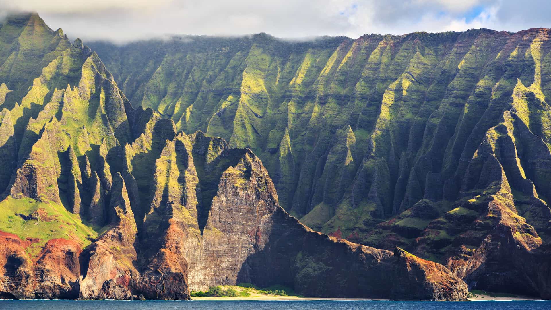 The dramatic and rugged Na Pali Coast on the island of Kauai, Hawaii. Massive, jagged green and brown cliffs with deep ridges and valleys rise steeply from the dark blue ocean. The lower parts of the cliffs are a reddish-brown color, while the upper parts are a lush, vibrant green. A small, secluded beach is visible at the base of the cliffs. The sky is partly cloudy, and the scene captures the iconic, awe-inspiring landscape of the Hawaii region.