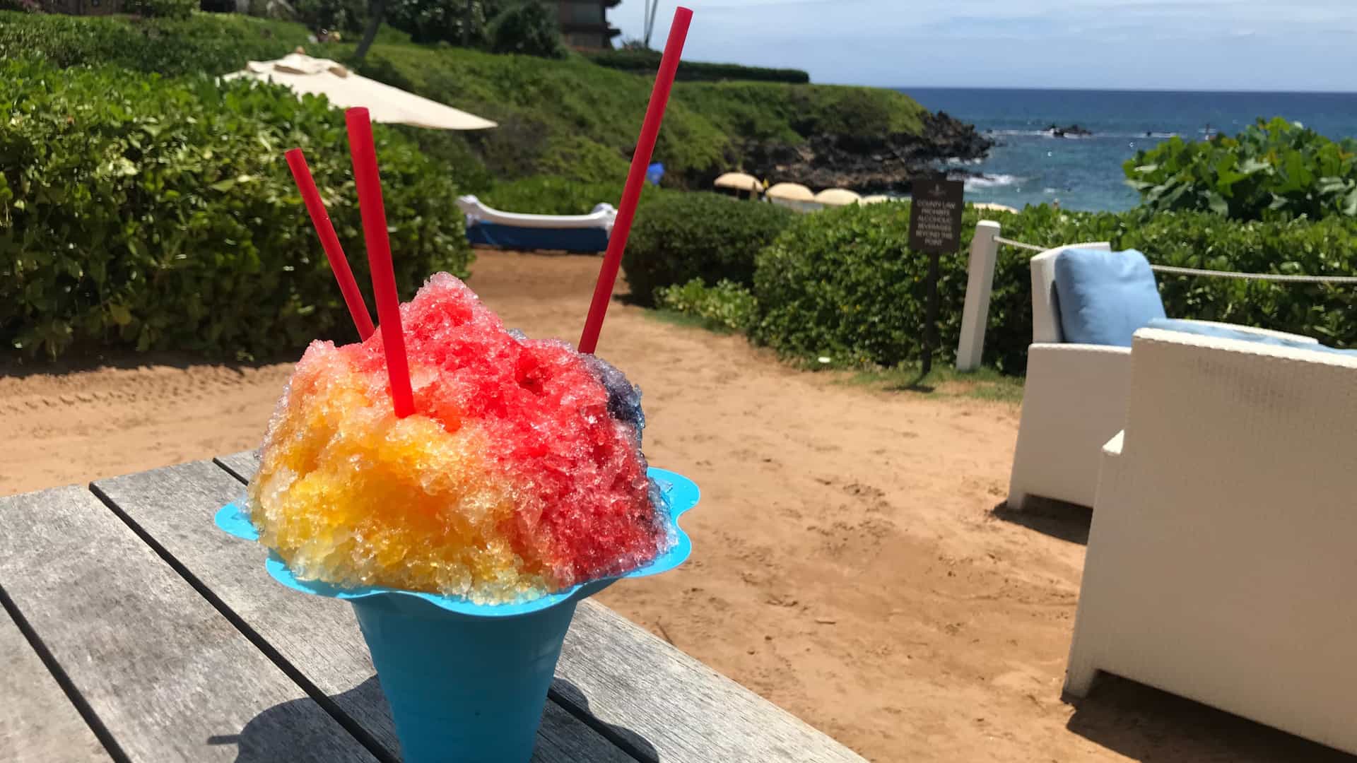 Hawaiian shaved iced in a shareable container on the beach with an ocean view in the background.