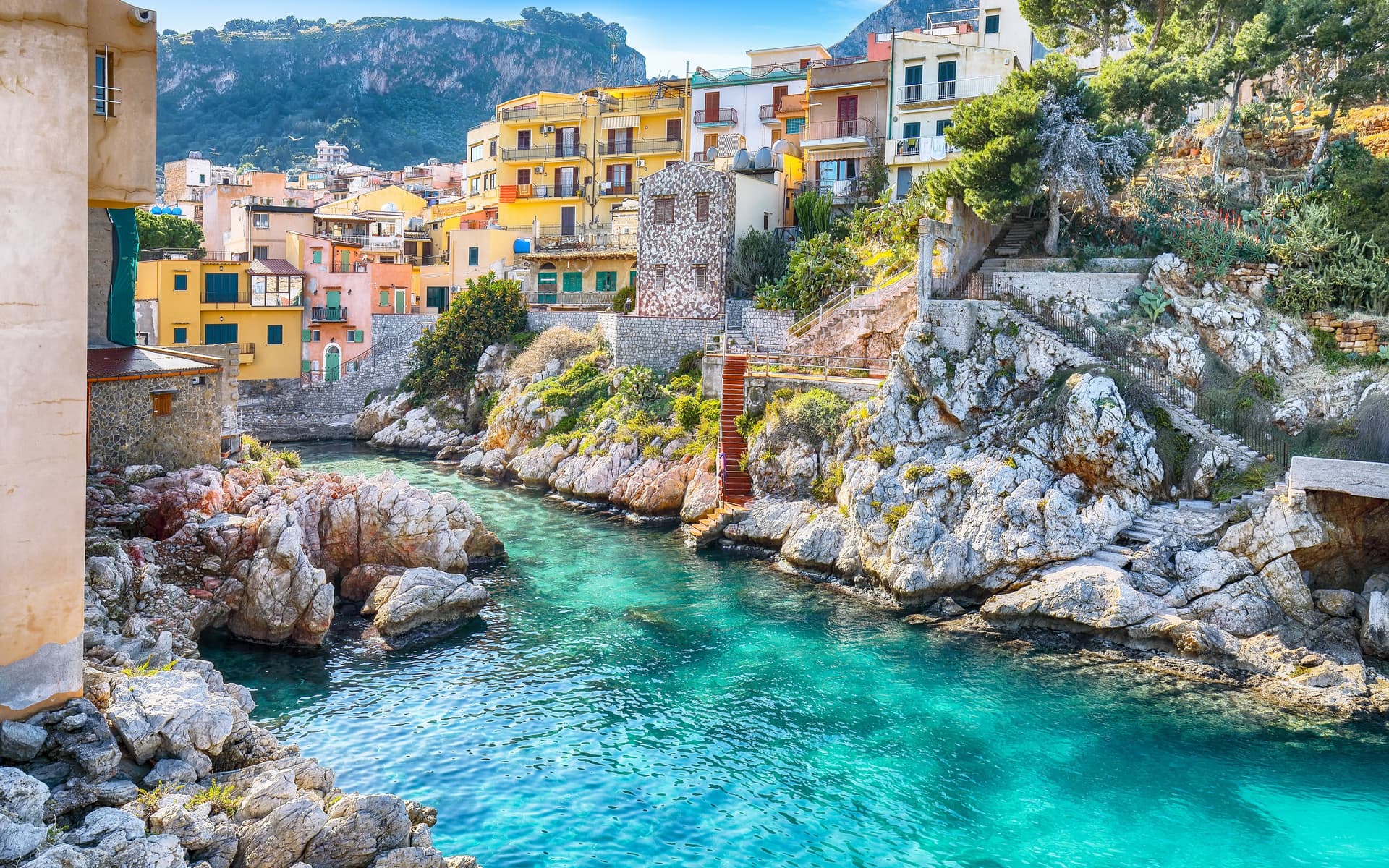 A vibrant coastal village with colorful yellow and pink buildings perched on rocky cliffs overlooking a narrow cove of crystal-clear turquoise water.