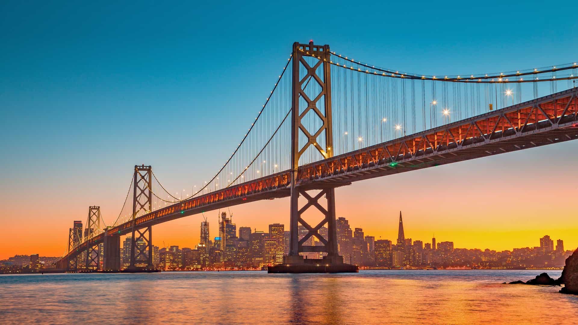 The San Francisco Bay Bridge and city skyline are beautifully lit at sunset, as seen from a Holland America cruise ship on a US Pacific Coast itinerary.