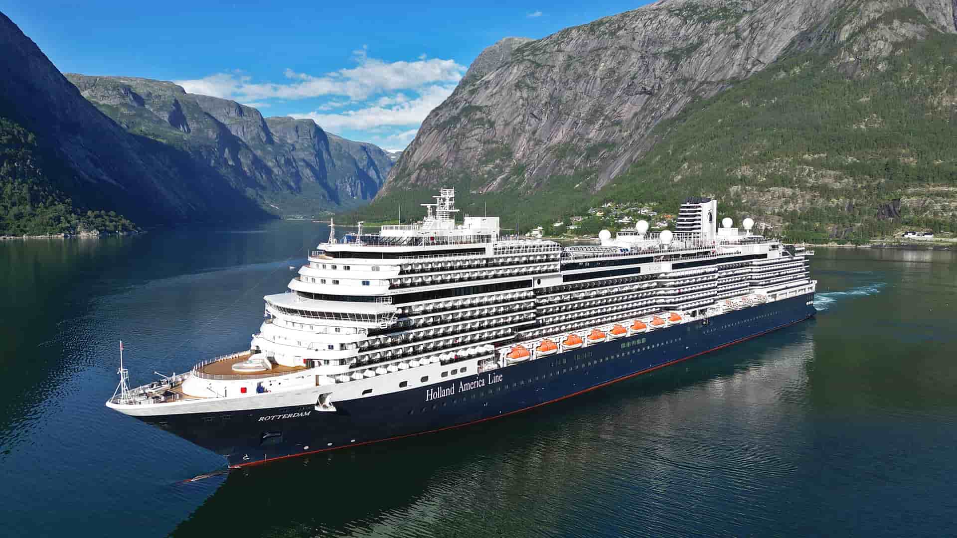 Holland America's Rotterdam at sea by a lush, green mountain.