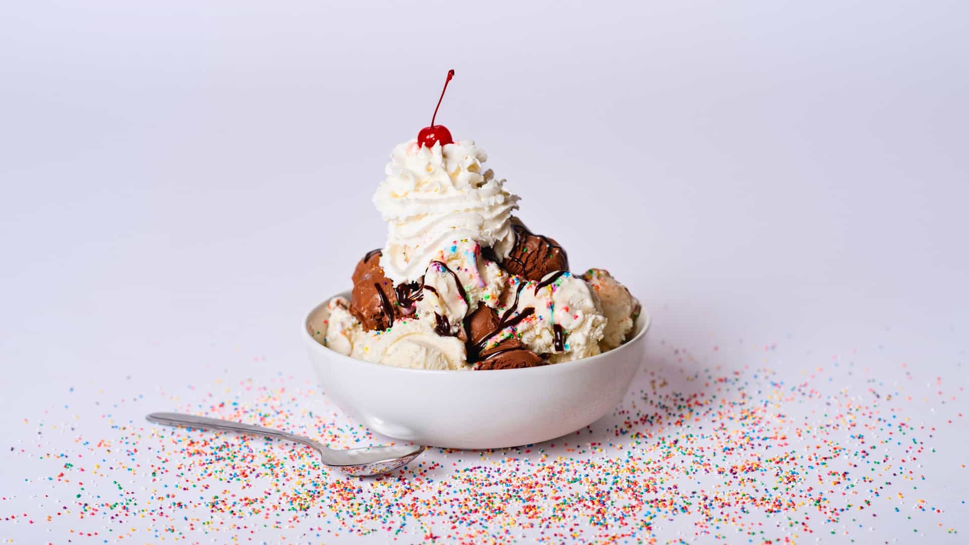 Ice cream sundae with a cherry on top and a plain background with a spoon and sprinkles scattered around the bowl.
