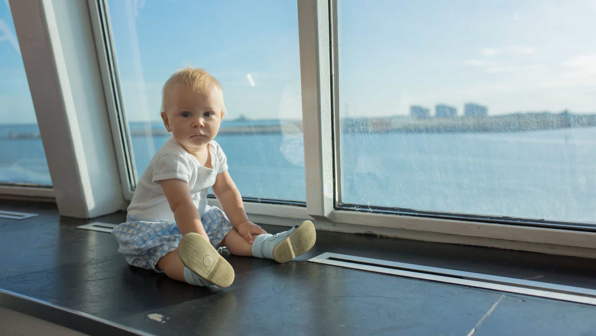A blonde baby sitting on a ledge inside a cruise ship, next to a large window at the blue ocean