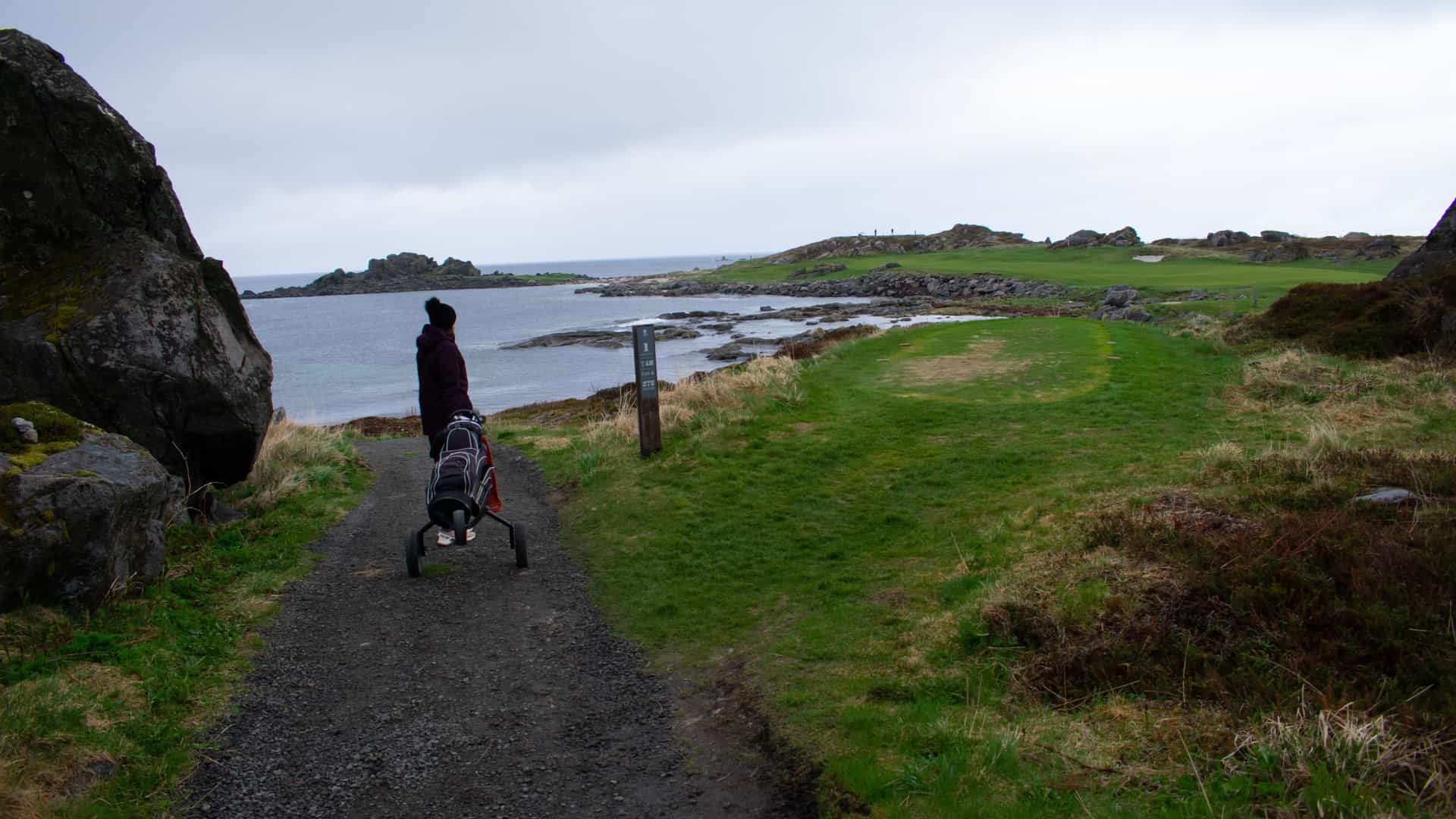 A golfer pulling a golf bag cart along a gravel path next to a rocky North Sea shoreline at Lofoten Links in Norway under an overcast sky.