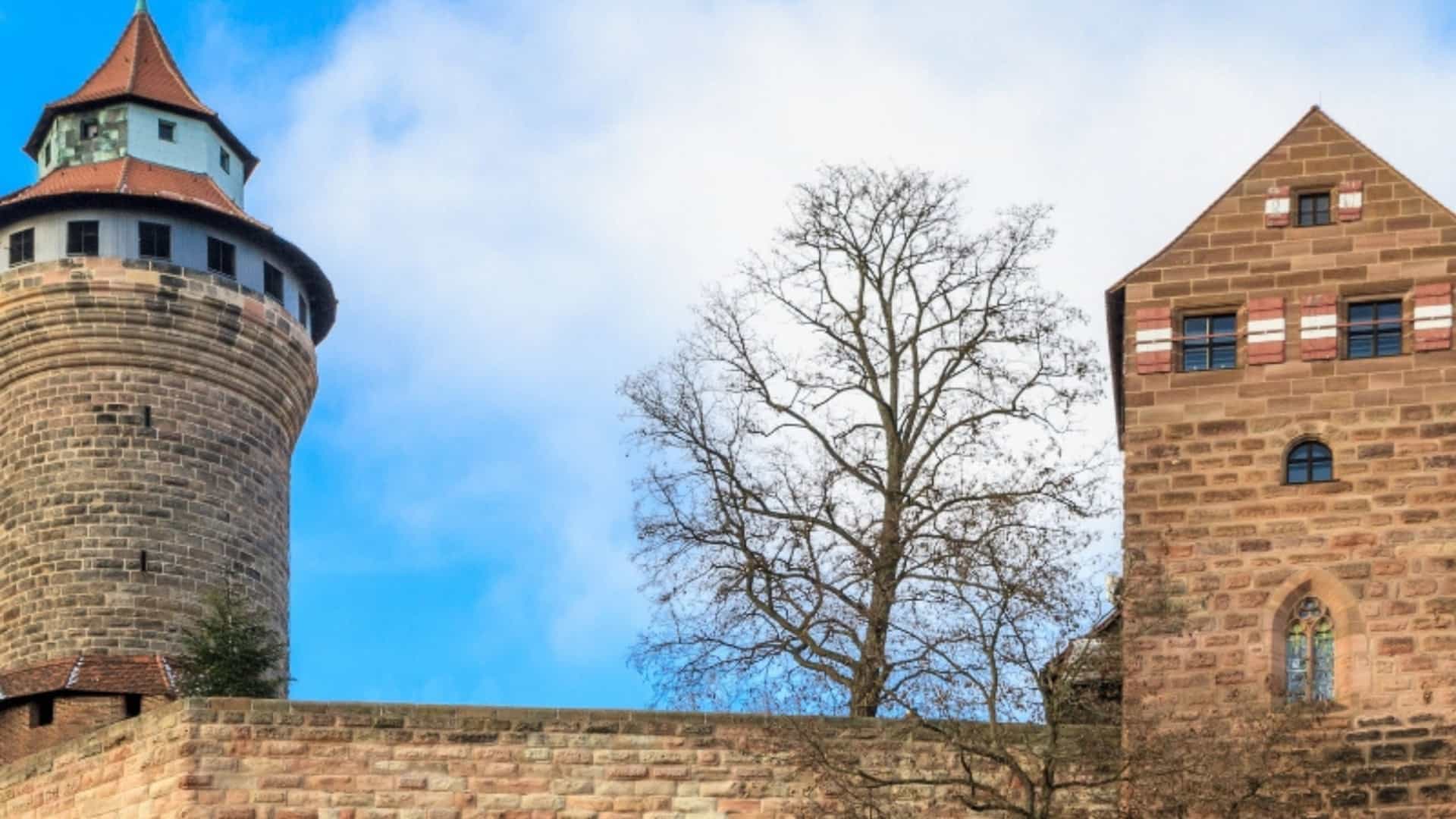 The towering, red-brick walls and a round turret of the Imperial Castle of Nuremberg, or Kaiserburg, in Germany. The stone walls are ancient and weathered. A bare tree stands in the courtyard between the two main structures. The sky is bright blue with scattered white clouds, highlighting the historic architecture of a city in the Main River region.