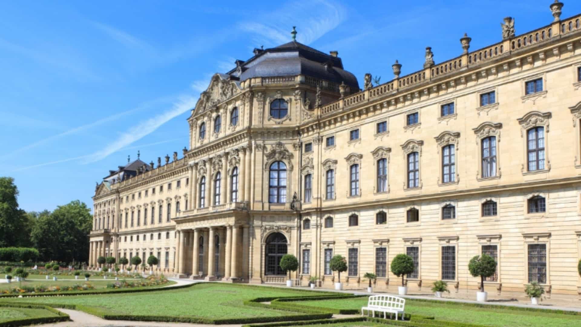 The grand facade of the Würzburg Residence, a stunning Baroque palace in Bavaria, Germany. The building is made of light-colored stone with rows of large windows and an ornate entrance. In front of the palace is a meticulously manicured garden with a geometric pattern of green hedges, gravel paths, and a small fountain. The sky is a brilliant blue with a few wispy clouds, highlighting the opulence and architectural splendor of a landmark in the Main River region.