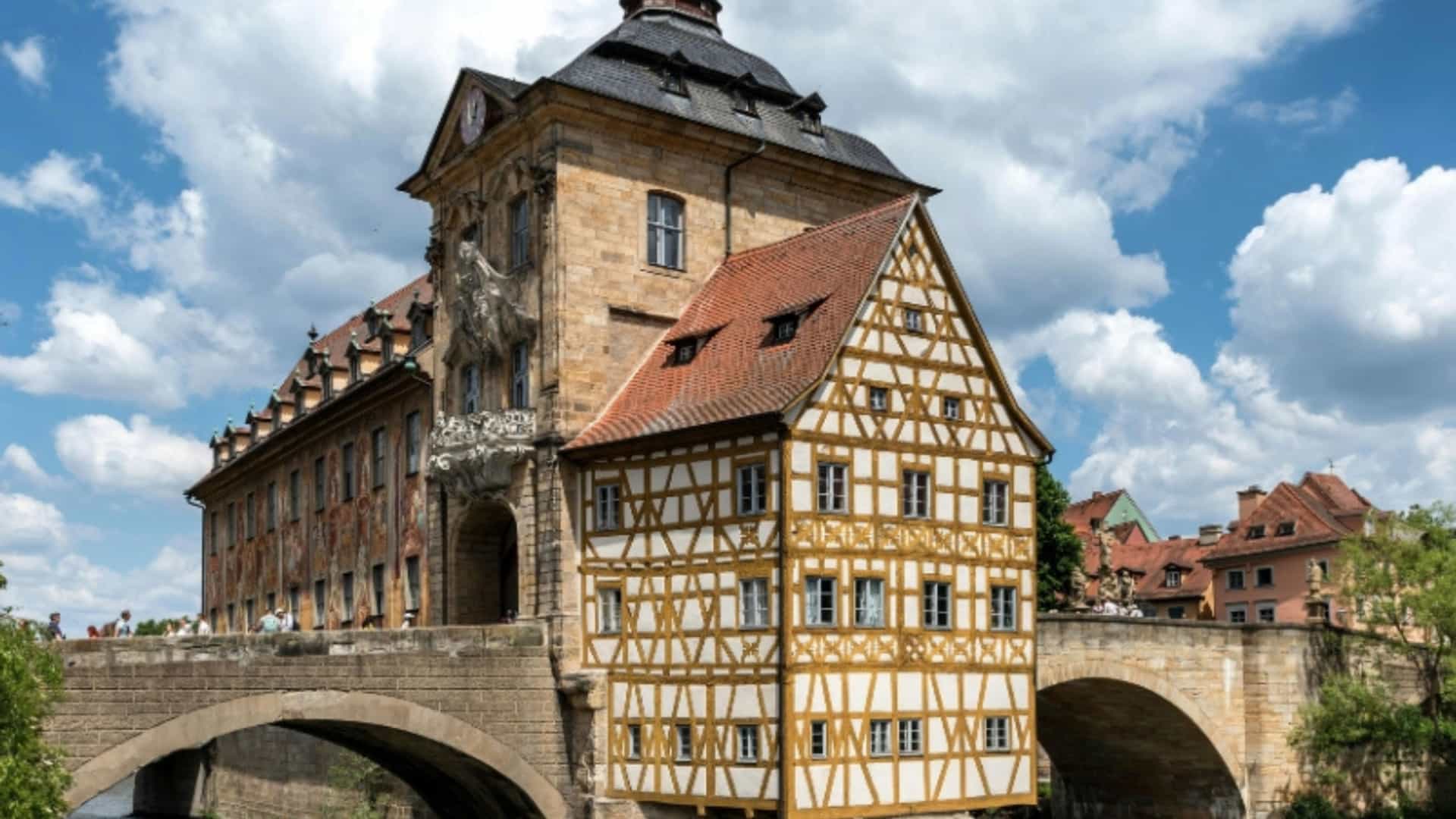A unique view of the Old Town Hall of Bamberg, Germany, built on an artificial island in the middle of the Regnitz River. The building has a mix of styles, including a stone section and a striking half-timbered facade with a white and yellow pattern. It is connected to the riverbanks by two stone arch bridges, with people crossing one of them. The sky is bright and filled with dramatic white clouds. The image captures the historical and picturesque nature of the city in the Main River region.