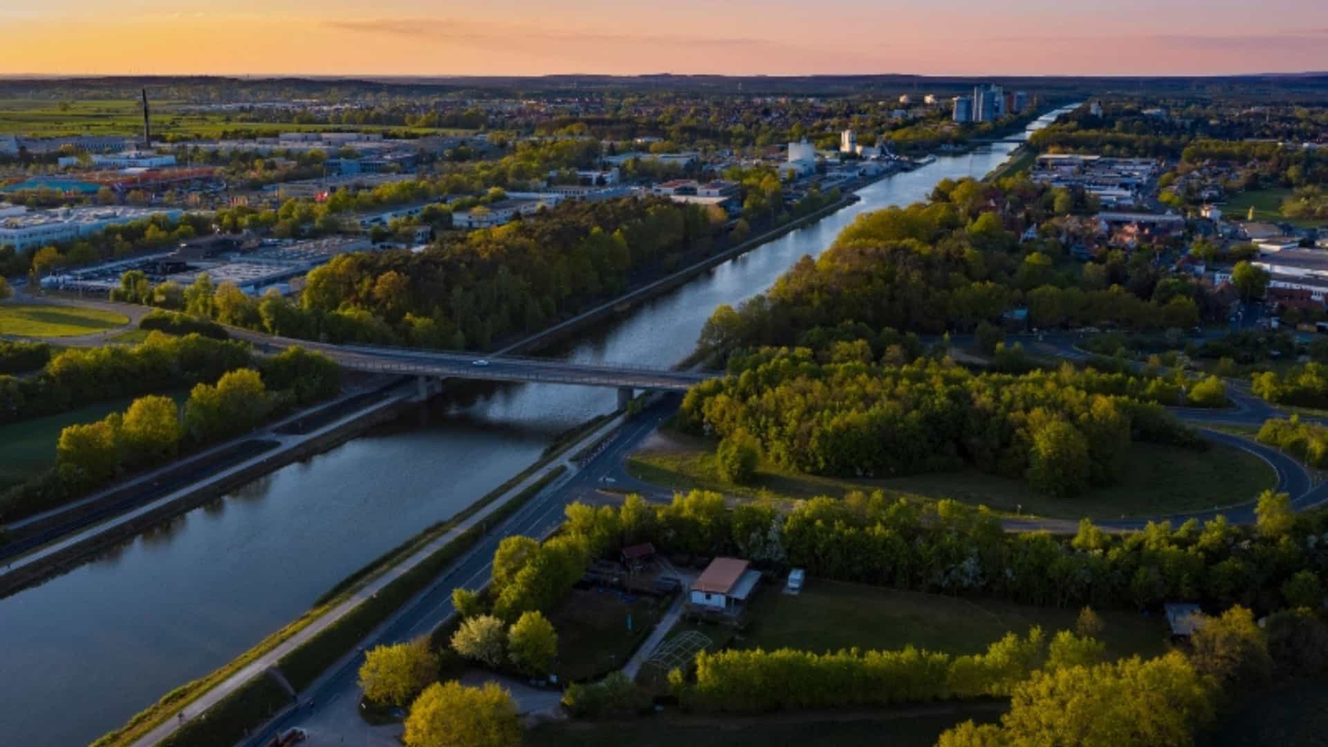 An aerial view of the Rhine-Main-Danube Canal in Germany at sunset. The canal snakes through a landscape of industrial buildings, residential areas, and green trees. A bridge crosses over the water, and roads curve alongside the canal and through the town. The sky is a gradient of deep blue to warm orange, casting a golden light over the scene. The image captures the confluence of man-made infrastructure and natural landscapes in this German region.