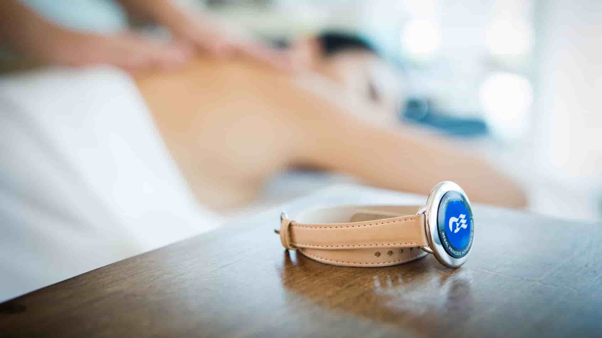 A close up shot of the Princess Cruises Medallion with a leather bracelet strap accessory, laid on a table, while there is a woman in the blurry background being massaged.