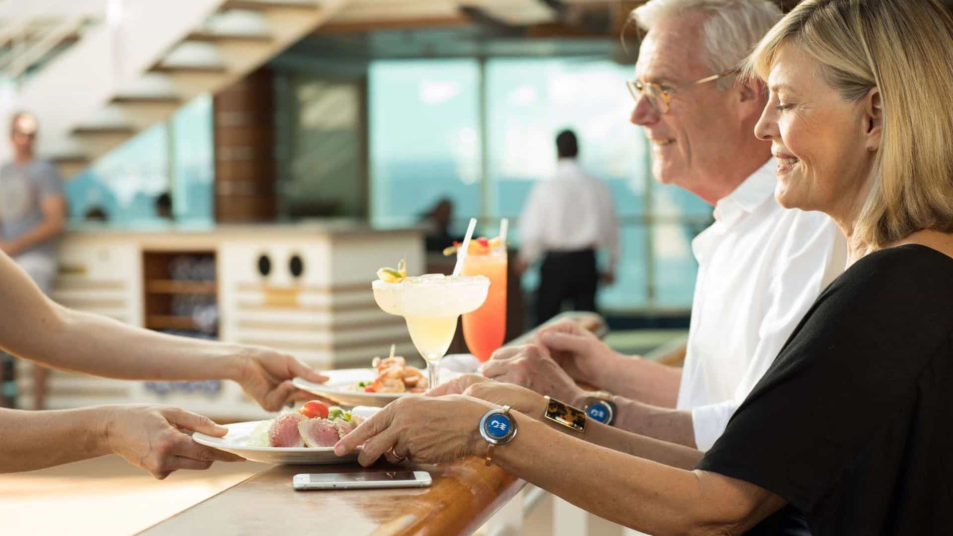 An older straight couple sit at by a railing aboard Regal Princess and order sushi & cocktails to their location using OceanNow through the Princess Cruises app.