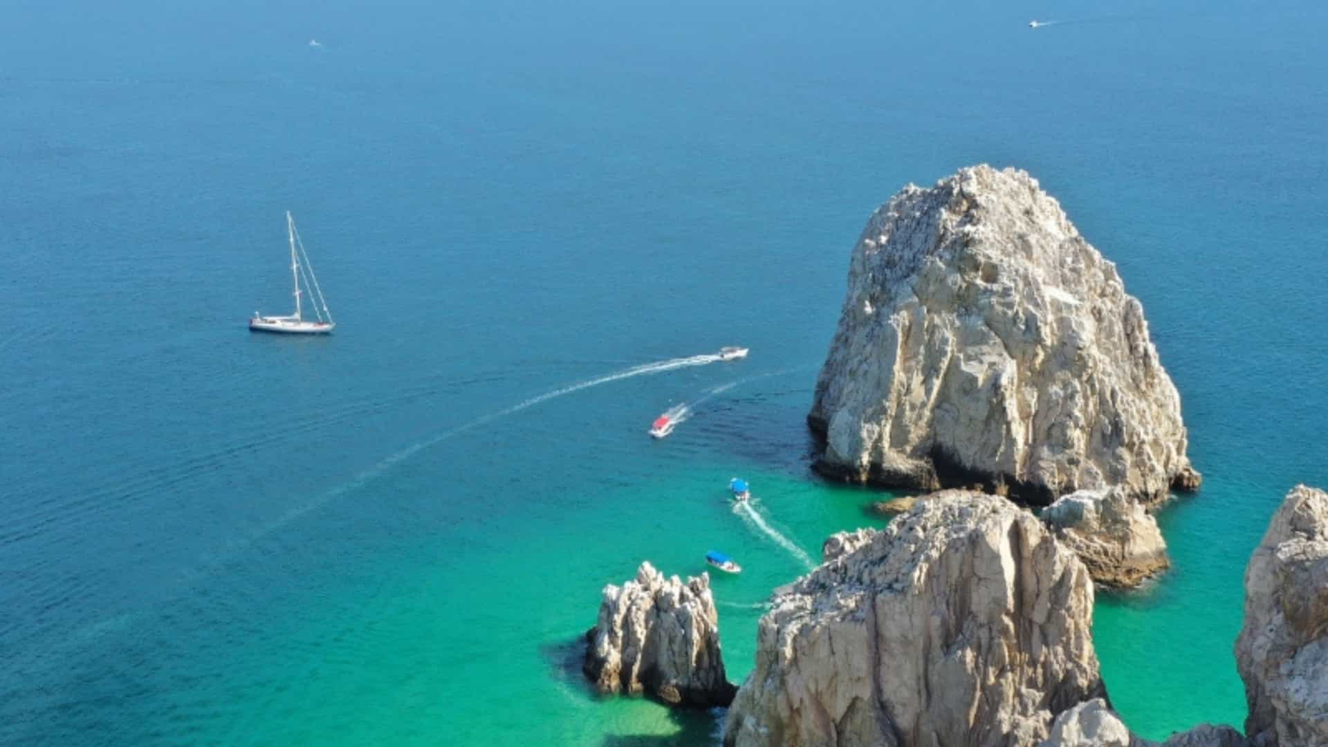 An aerial view of the iconic rock formations and turquoise waters of Cabo San Lucas, Mexico. Several boats, including a white sailboat, speedboats, and parasailers, navigate the clear blue water. The massive, jagged rock formations rise from the sea, creating a stunning and dramatic coastal landscape. The image captures the popular boating and water-sport activities in this beautiful Mexican region.