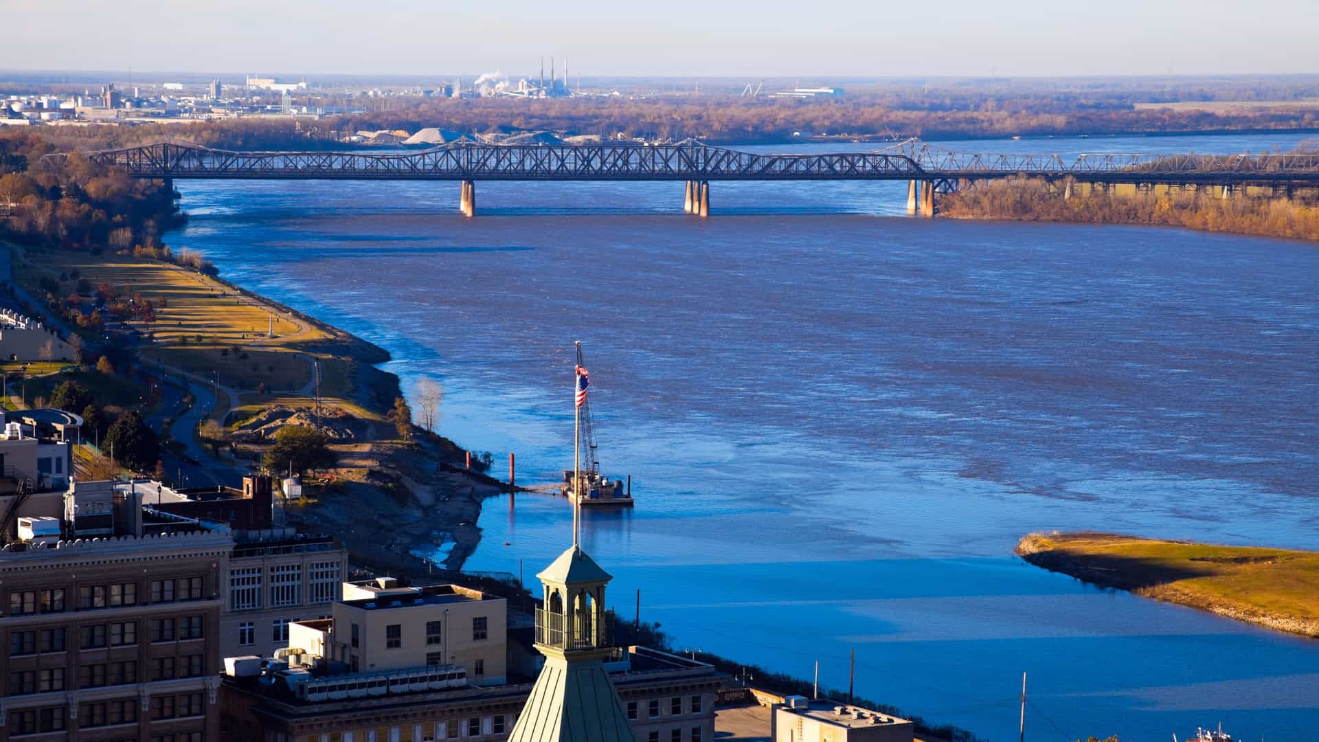 An elevated view of the Mississippi River from Memphis, Tennessee. The wide, blue river flows under a large steel bridge. On the left bank, the cityscape of downtown Memphis is visible, with buildings in the foreground and a park area along the water's edge. The right bank is a mix of brown and green trees and an industrial area. The sky is bright and clear, showcasing the bustling and expansive landscape of this significant American river.