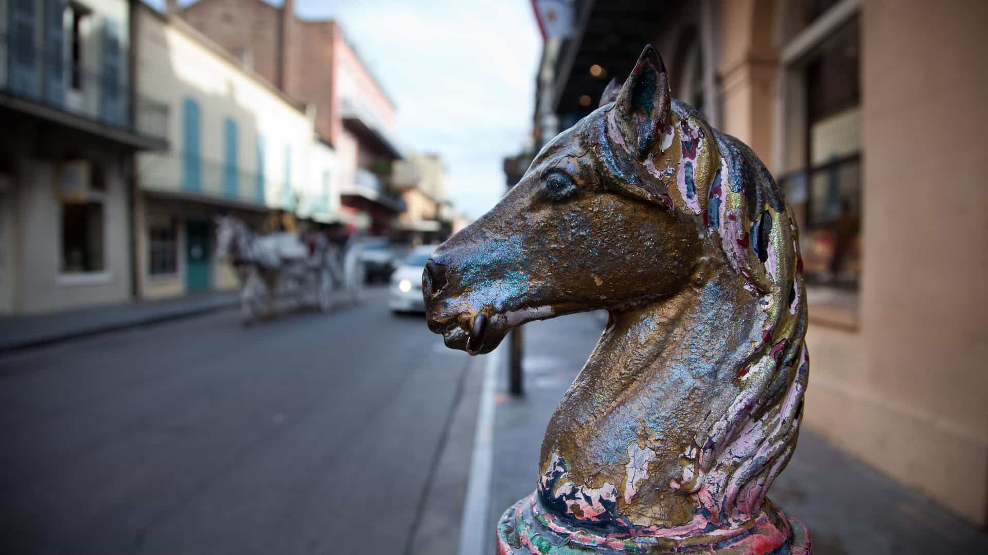 A low-angle, close-up shot of a colorful, weathered horse head sculpture atop a street bollard in the French Quarter of New Orleans, Louisiana. The metal horse head is painted in shades of gold, blue, and red, with visible peeling paint. In the background, a street in the French Quarter is visible, with blurred historic buildings, a horse-drawn carriage, and a car. The image captures the unique, artistic, and historical atmosphere of this iconic Mississippi River city.