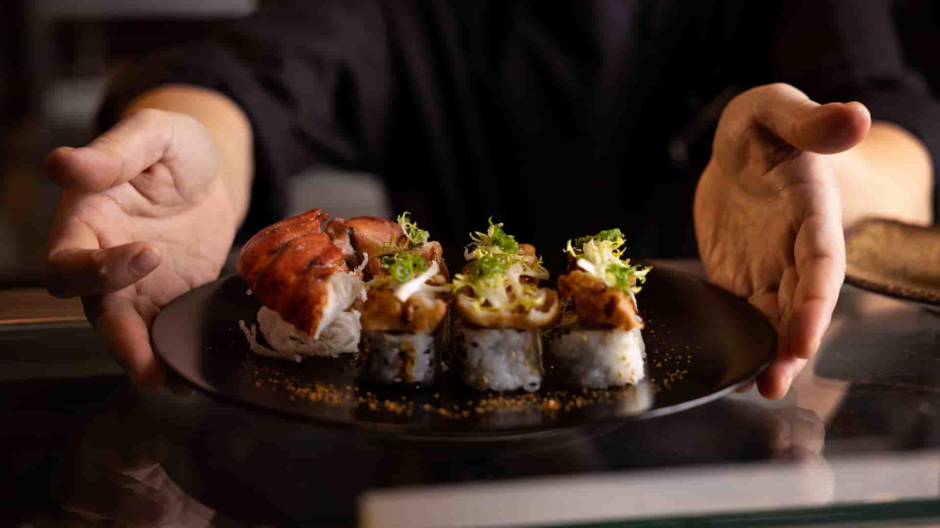 A close up shot of tempura lobster sushi roll from Nami Sushi aboard Holland America Line. The plate of sushi is being held by a chef's hands in black uniform and slightly tilted forward to show the camera it's delicate plating.
