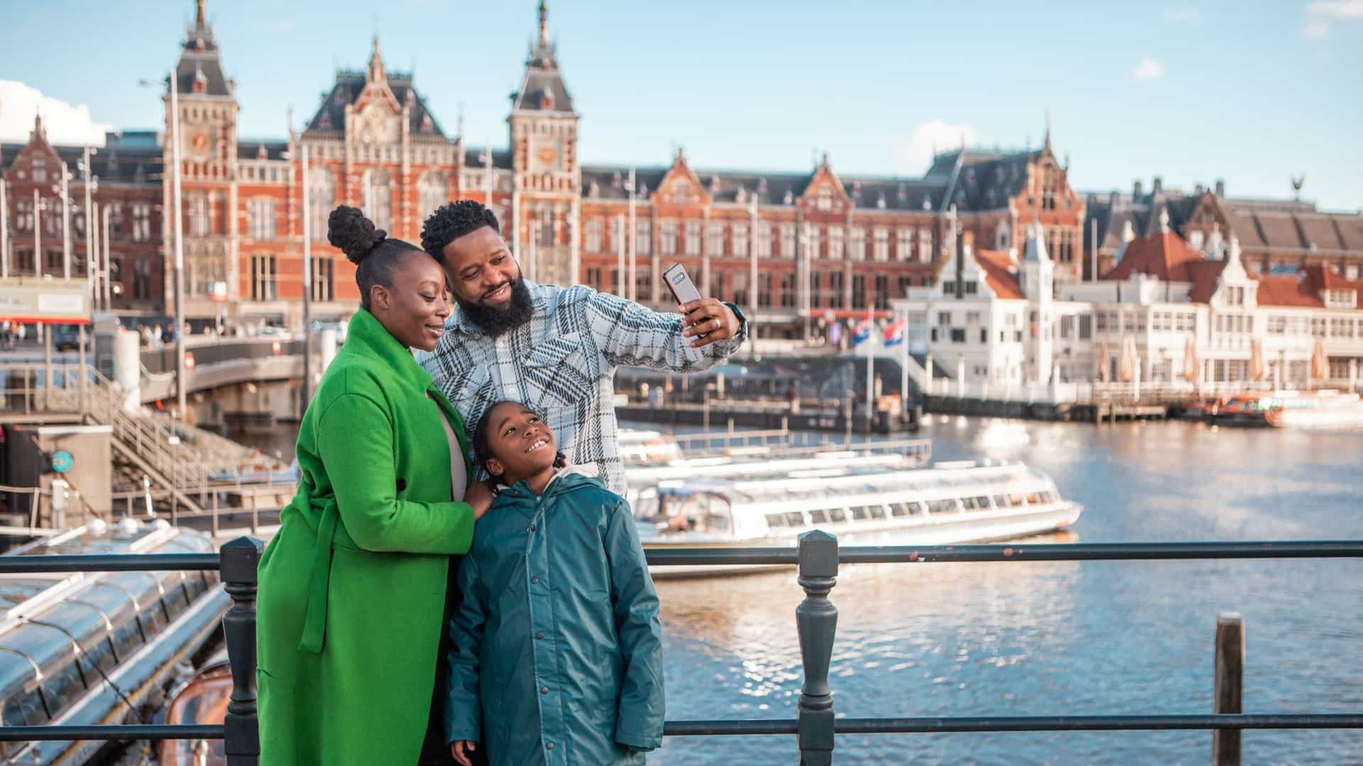 A Black family of three takes a selfie on a bridge in Amsterdam, Netherlands, with a historic building and canal in the background. The father holds the phone, while the mother in a green coat and their child smile.