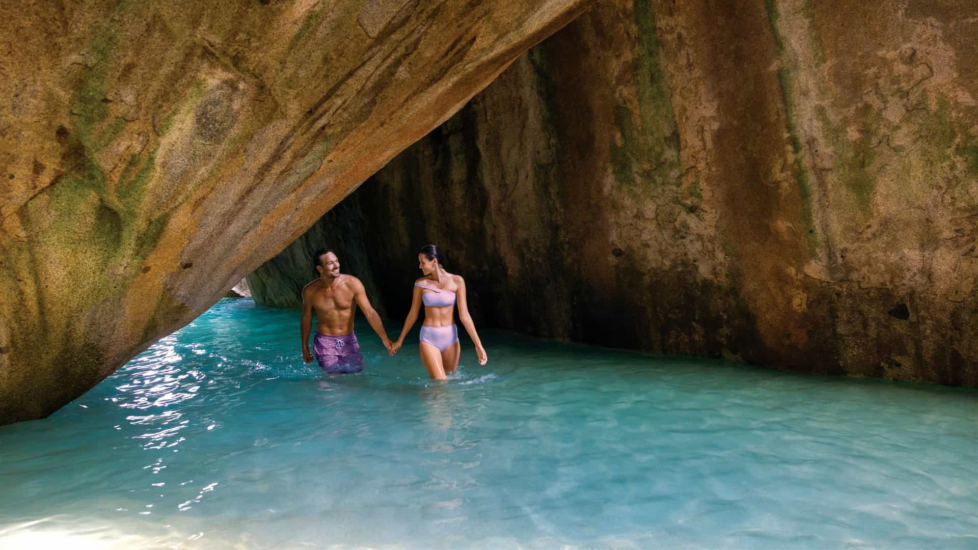 A tanned couple, a man in purple swim trunks and a woman in a light purple bikini, walk hand-in-hand through the Virgin Gorda Baths, which are a geological wonder located on the island of Virgin Gorda in the British Virgin Islands. They are characterized by massive granite boulders that form unique caves, grottos, and tidal pools, creating a beautiful and adventurous landscape.