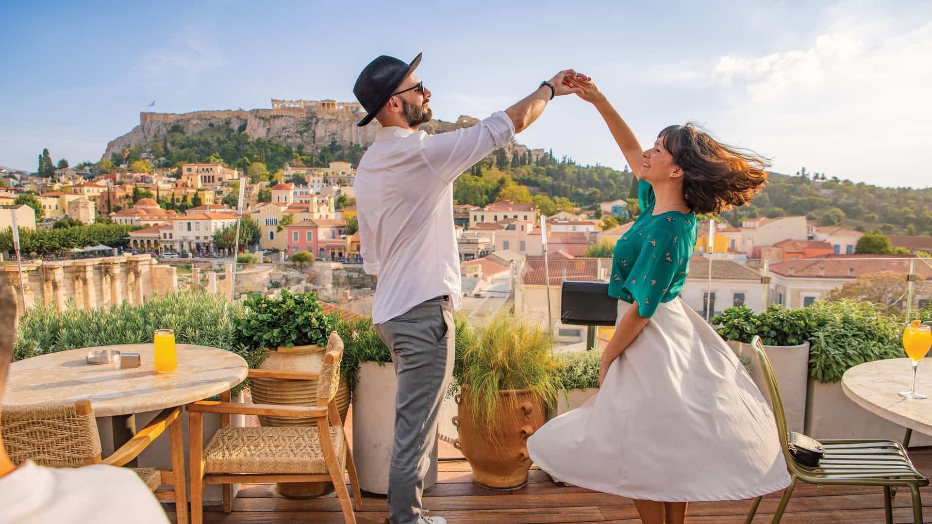 A smiling couple dances on a rooftop terrace in Athens, Greece, with a scenic view of the city and the historic Acropolis in the background.