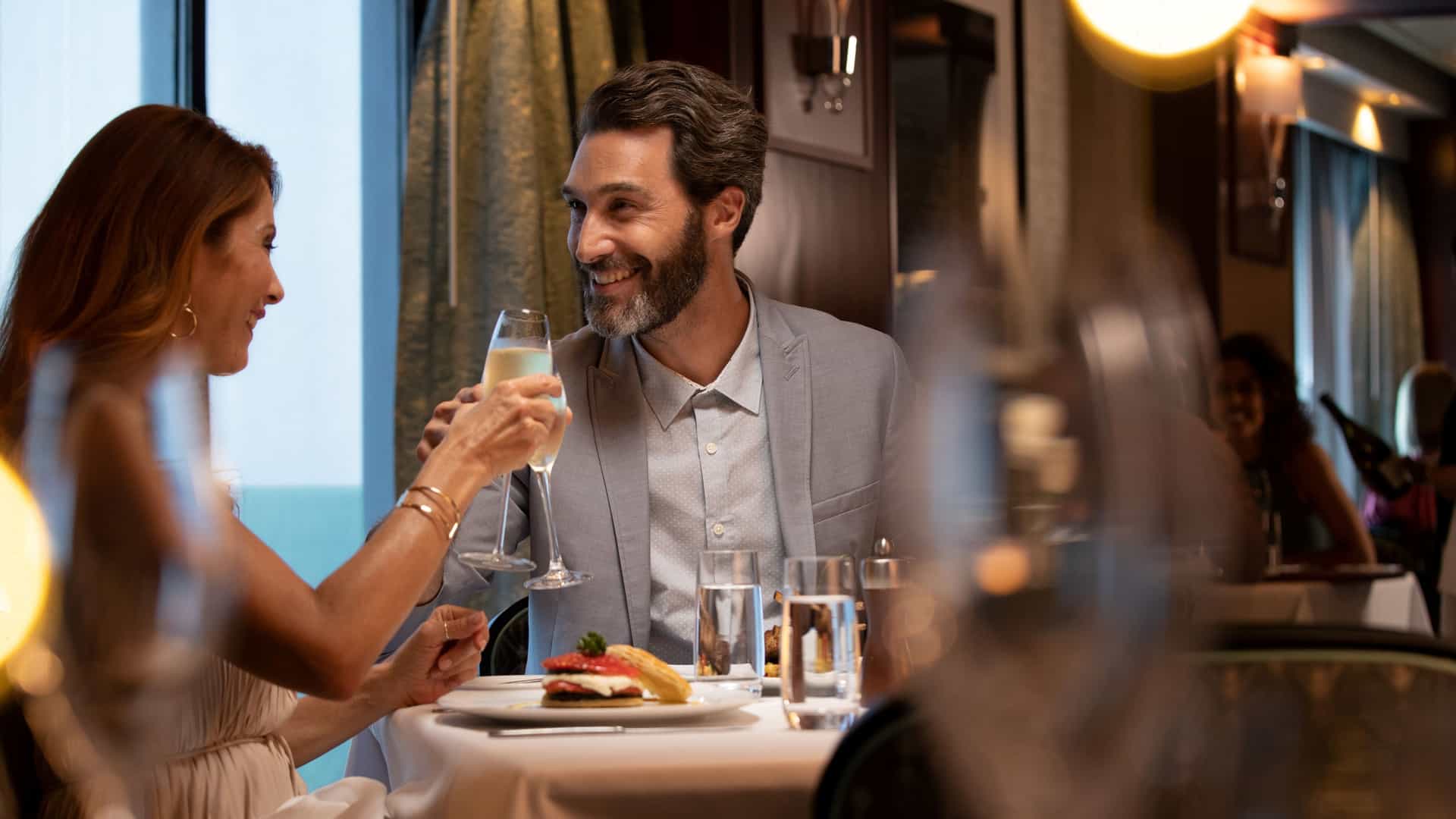A smiling couple toasts with champagne glasses while dining at a white tablecloth table inside a specialty restaurant on a Norwegian Cruise Line ship.
