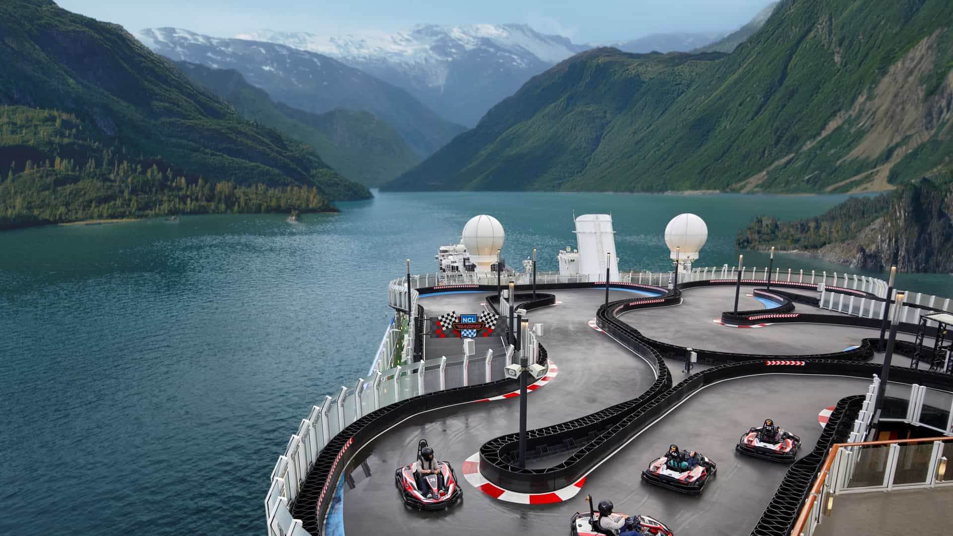 A thrilling go-kart racetrack is shown on the top deck of the Norwegian Bliss cruise ship, with go-karts racing along the track. In the background is a scenic view of an Alaskan fjord with green mountains and snow-capped peaks.
