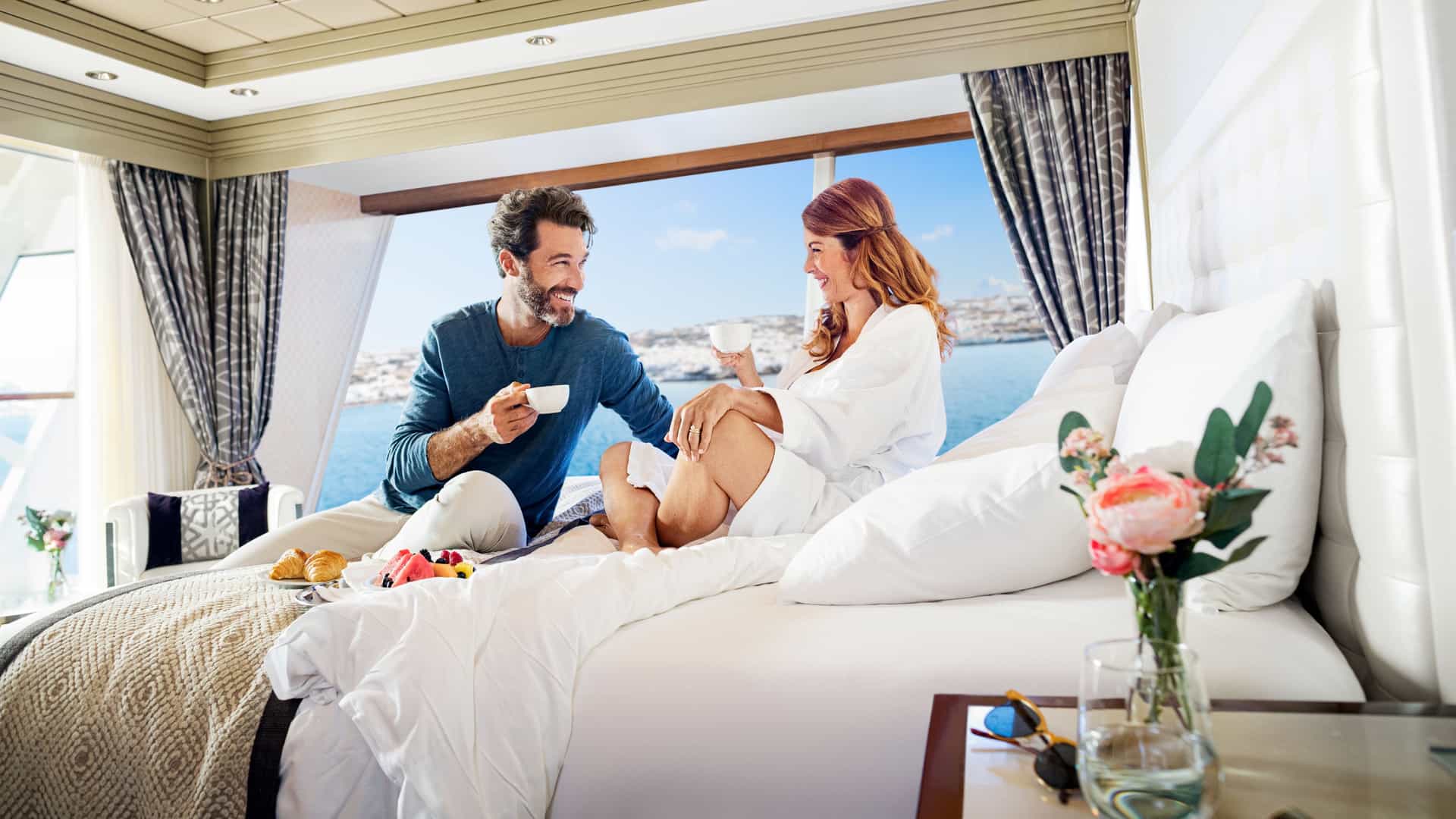 A smiling couple enjoys breakfast in bed with coffee and fresh fruit inside a luxury cruise ship suite featuring expansive floor-to-ceiling windows that overlook the blue ocean.
